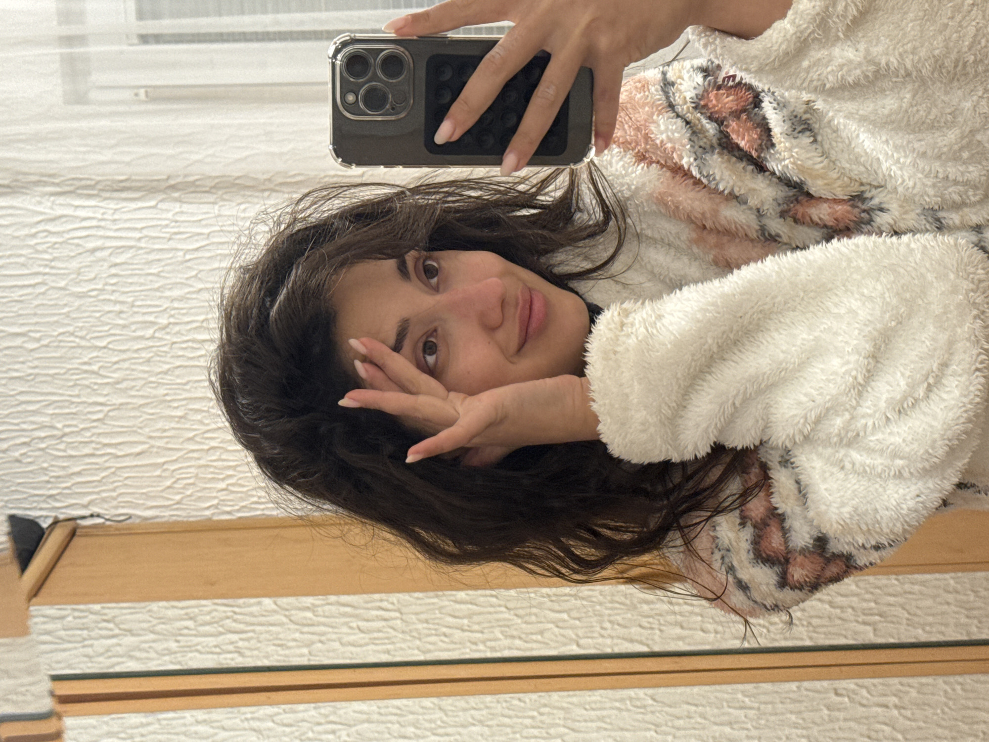 A young woman is taking a selfie in a mirror. She is wearing a soft, patterned robe and holding a smartphone. The reflection in the mirror shows a textured wall and a window. The image suggests an indoor setting, likely a personal residence. The city or location is not evident from visual details alone.