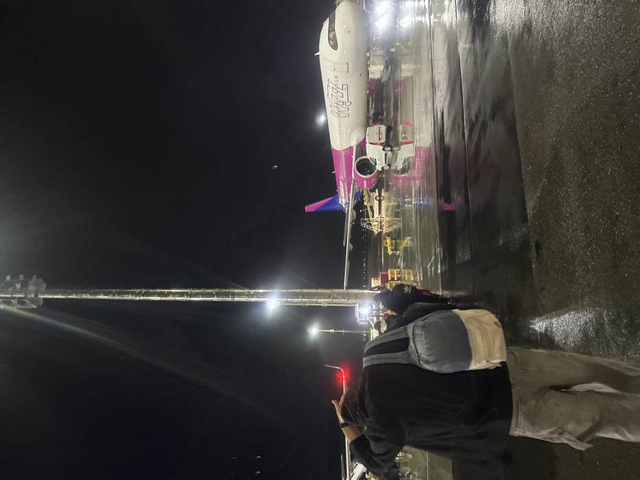 An airplane, likely a Wizz Air plane, is parked at an airport at night. The ground appears wet, possibly from rain. A pedestrian bridge extends towards the plane. A person stands in the foreground, facing the aircraft and possibly taking a photo of it. The scene is illuminated by airport lights.
