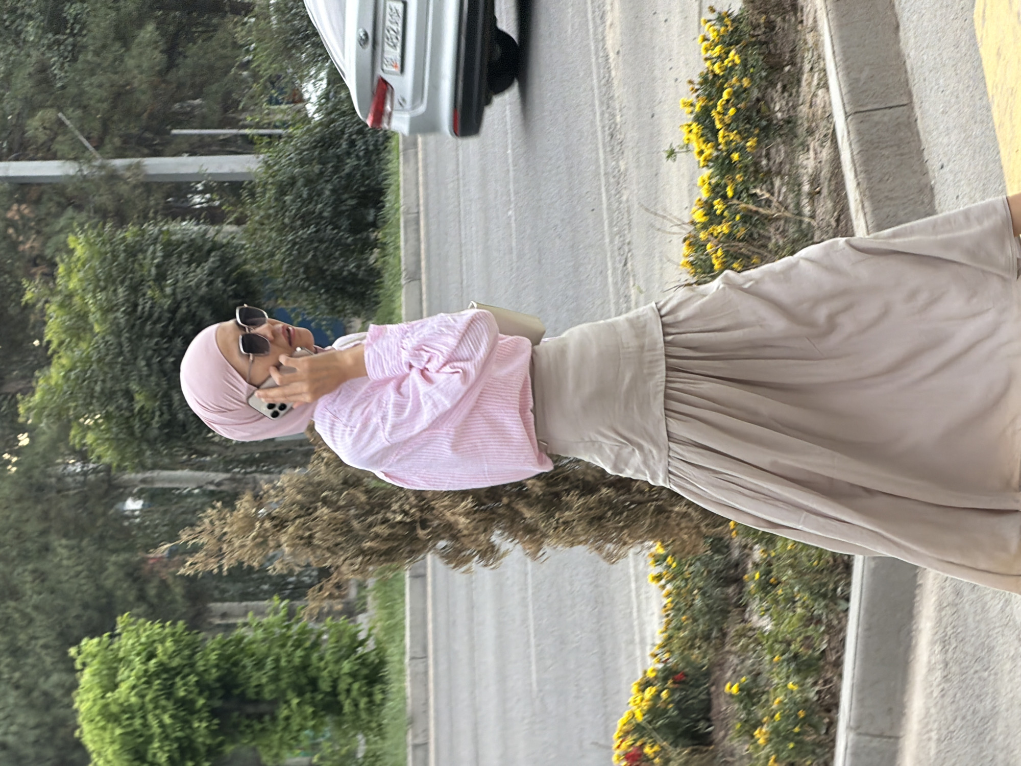 A woman wearing a pink hijab, sunglasses, and a pink striped shirt is walking and talking on her phone. She is also wearing a beige maxi skirt and carrying a light-colored purse. The woman is walking on a sidewalk next to a road with a silver car driving by. There are trees and yellow flowers in the background.