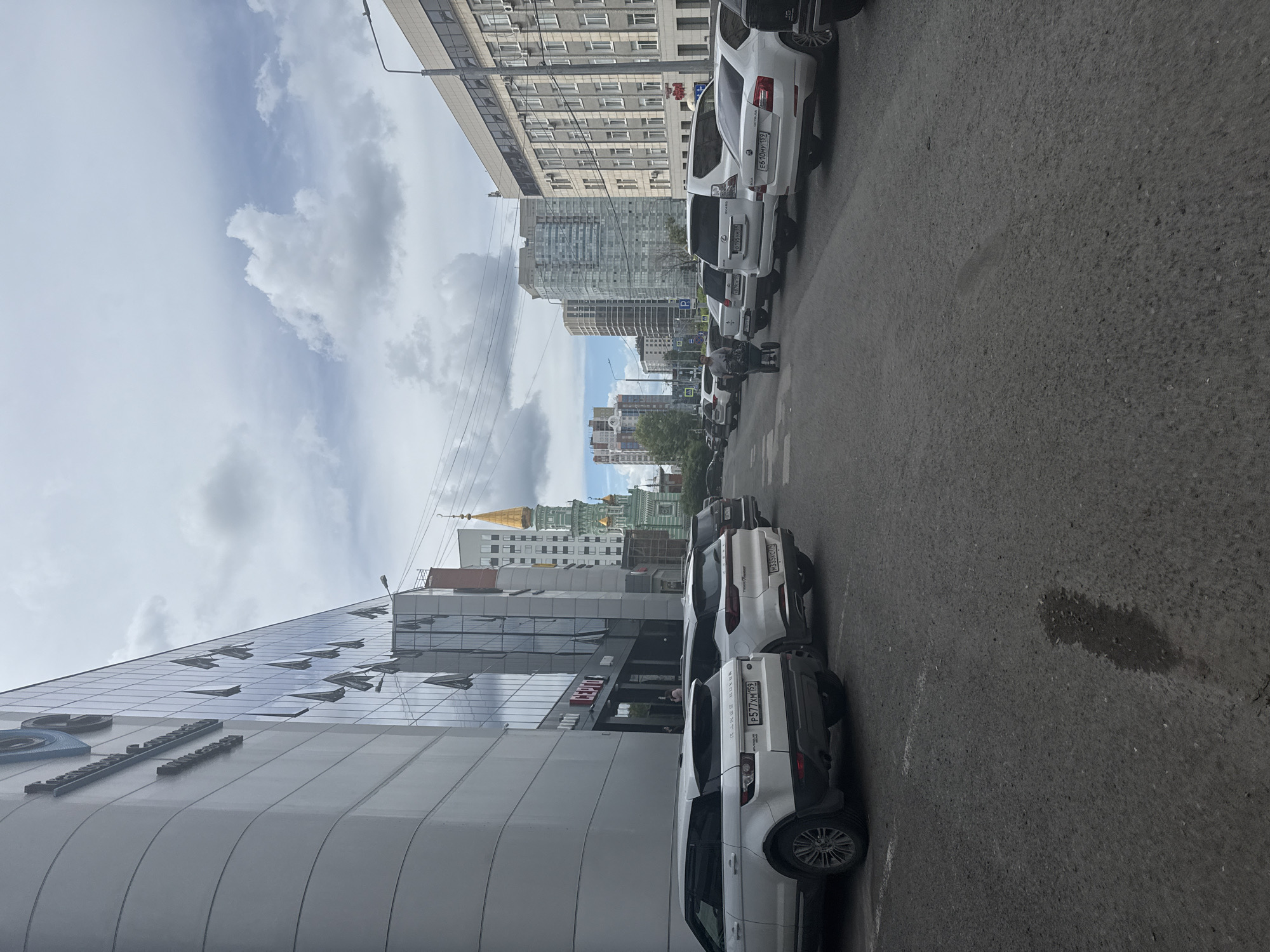 The image depicts a street scene in Perm, Russia, with a large modern building on the left and several parked cars on the right. The building on the left features a curved, grey facade with horizontal banding and a reflective glass section with many open windows. The word "СЕРГТО" is visible on a sign attached to the building. In the background, a distinctive green and gold domed tower rises above other buildings. A row of parked cars, including white SUVs and sedans, lines the street. A person is pushing a wheelbarrow down the street. The sky is overcast with scattered clouds. The street is paved with asphalt and shows signs of wear, including a wet patch on the ground.