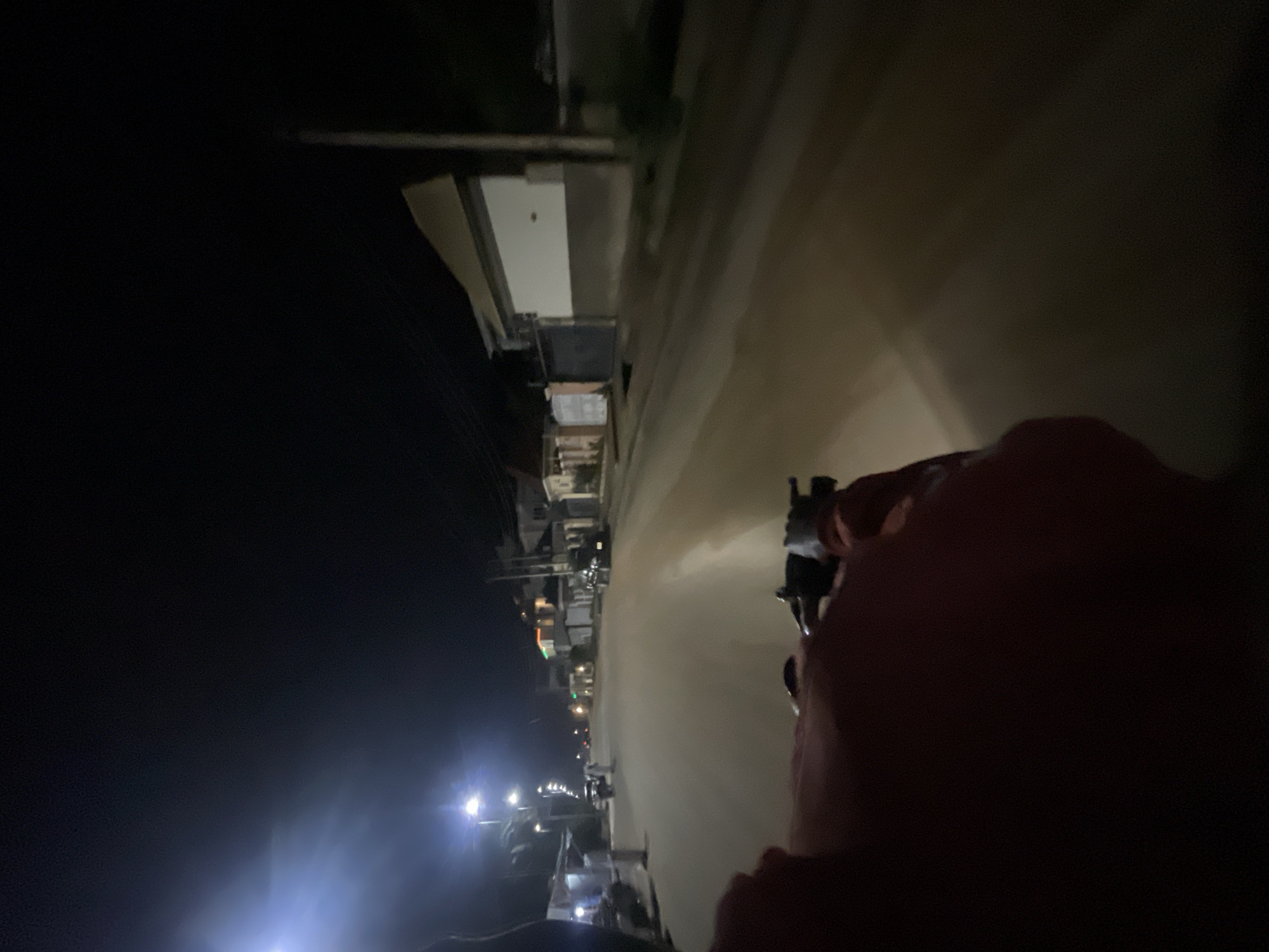 The image, rotated 90 degrees counter-clockwise, depicts a first-person perspective from a vehicle, likely a motorcycle or scooter, traveling down an urban street at night. In the immediate foreground on the right, a person's blurred hand or arm, possibly wearing a reddish sleeve, grips a dark handlebar.
The street, which appears to be paved, is the central focus, illuminated by a strong, elongated path of light extending into the distance. This illumination stems from a combination of the vehicle's headlights and several bright streetlights positioned along the left side of the road. On the left, a series of low-rise buildings and structures line the street, their forms partly visible against the dim night and the scattered light. Some structures show faint internal lighting or architectural details. In the far distance, bright, almost blinding, headlights of an approaching or following vehicle are visible. The sky above is dark, confirming the nighttime setting.
The image conveys a sense of motion and nocturnal travel through a bustling, yet dimly lit, urban environment. The strong contrast between the dark surroundings and the intense artificial light sources is a prominent visual feature. The general appearance of the street and buildings suggests an urban area, consistent with the provided context of Abuja, Nigeria. No discernible text is visible in the image.