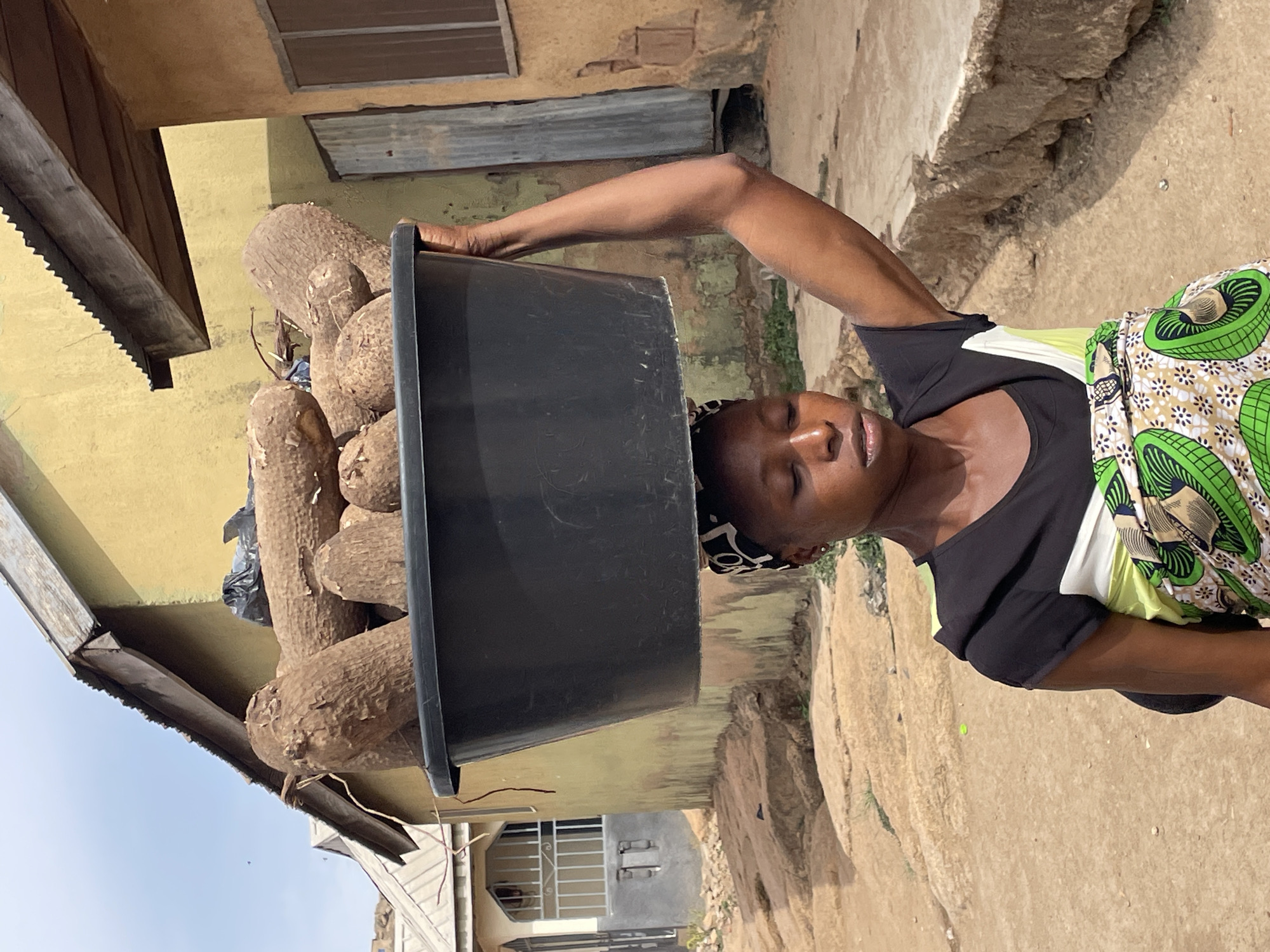 A young woman is depicted outdoors, balancing a large, dark-colored basin filled with numerous yams on her head. Her body is angled, and her right arm is raised, possibly to steady the heavy load. She wears a black t-shirt adorned with a light green and white patterned detail on the side, and a brightly colored, patterned wrap skirt featuring green, white, and brown elements. Her eyes appear to be closed or looking downwards, suggesting concentration or effort. A dark, patterned headscarf or bandana is visible around her forehead.
The background shows parts of simple buildings with ochre or yellow-painted walls, a window with a metal frame, and sections of corrugated metal roofing. A wooden beam with a distinctive serrated edge is visible in the upper left corner, possibly part of a roof structure. The ground is dry, light-colored earth or concrete, appearing somewhat uneven. The sky in the upper left is clear and light blue, indicating bright daylight and sunny weather. There are no visible signs of text. The scene conveys a moment of daily life, likely in a residential or market area in Jos, Nigeria, where head-carrying is a traditional method of transporting goods.