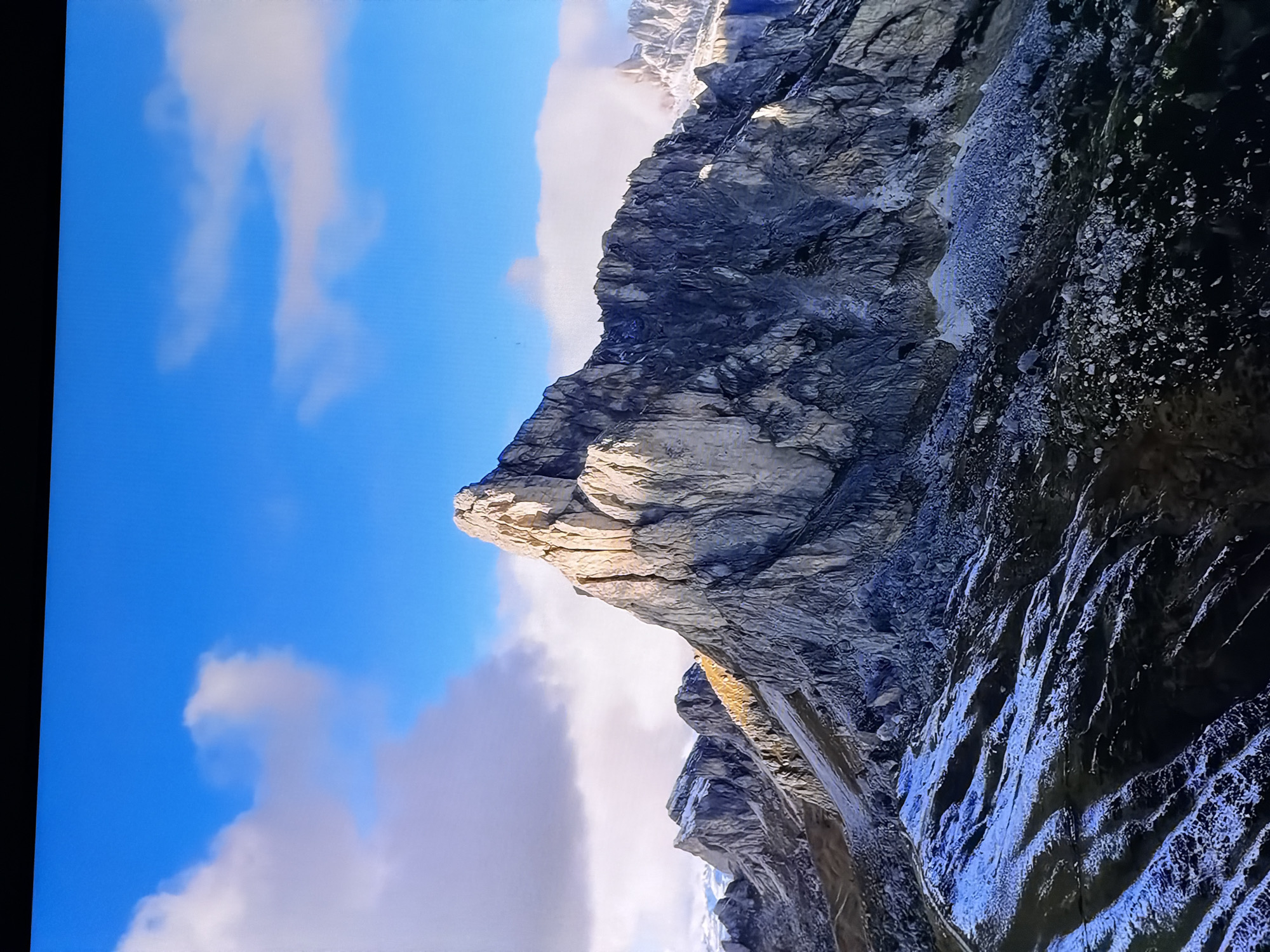 The image displays a dramatic mountain landscape under a vibrant blue sky with scattered clouds. The foreground is a rugged, rocky terrain with patches of snow, suggesting a high-altitude or early winter environment. Several imposing, jagged mountain peaks dominate the midground and background, characterized by their steep, rocky faces and sharp ridges. Sunlight illuminates parts of the mountains, creating strong contrasts between light and shadow. The overall scene is one of natural grandeur and remote wilderness. This image is not located in Chirchik, Uzbekistan, as the scenery is clearly mountainous and does not depict urban or any recognizable features of that city.