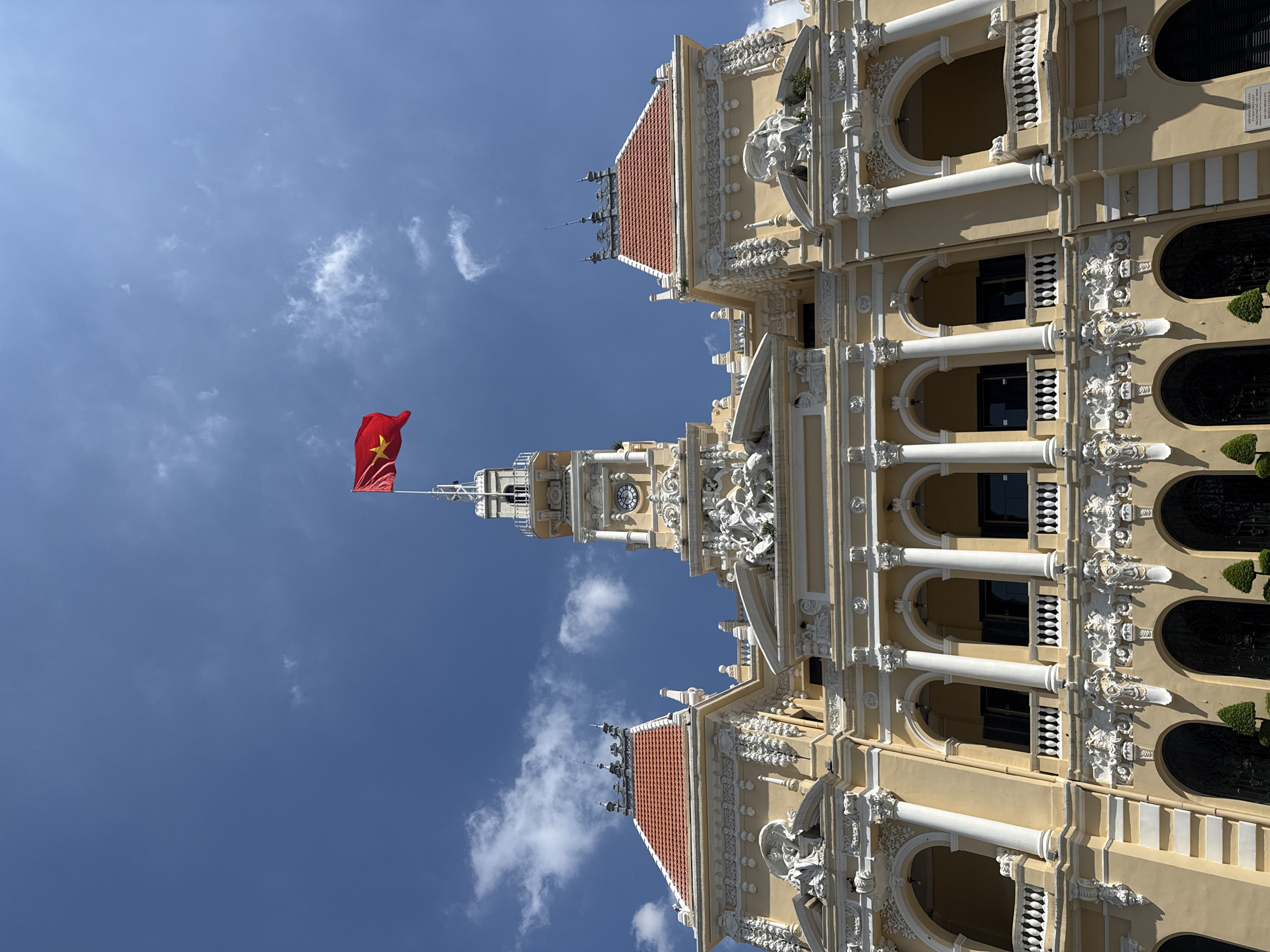 A low-angle, full shot captures the exterior of the Ho Chi Minh City Hall, also known as the People's Committee Building, in Thủ Đức, Vietnam. The imposing, colonial-era structure is painted a pale yellow with intricate white decorative elements. A prominent clock tower rises from the center of the building, topped by a flagpole displaying the Vietnamese flag, a red flag with a yellow star. The building features multiple arched windows and balconies supported by columns, with ornate sculptures adorning the facade. Red-tiled roofs cap the structure. The sky above is a clear blue, with scattered white clouds. No people are visible in the frame.