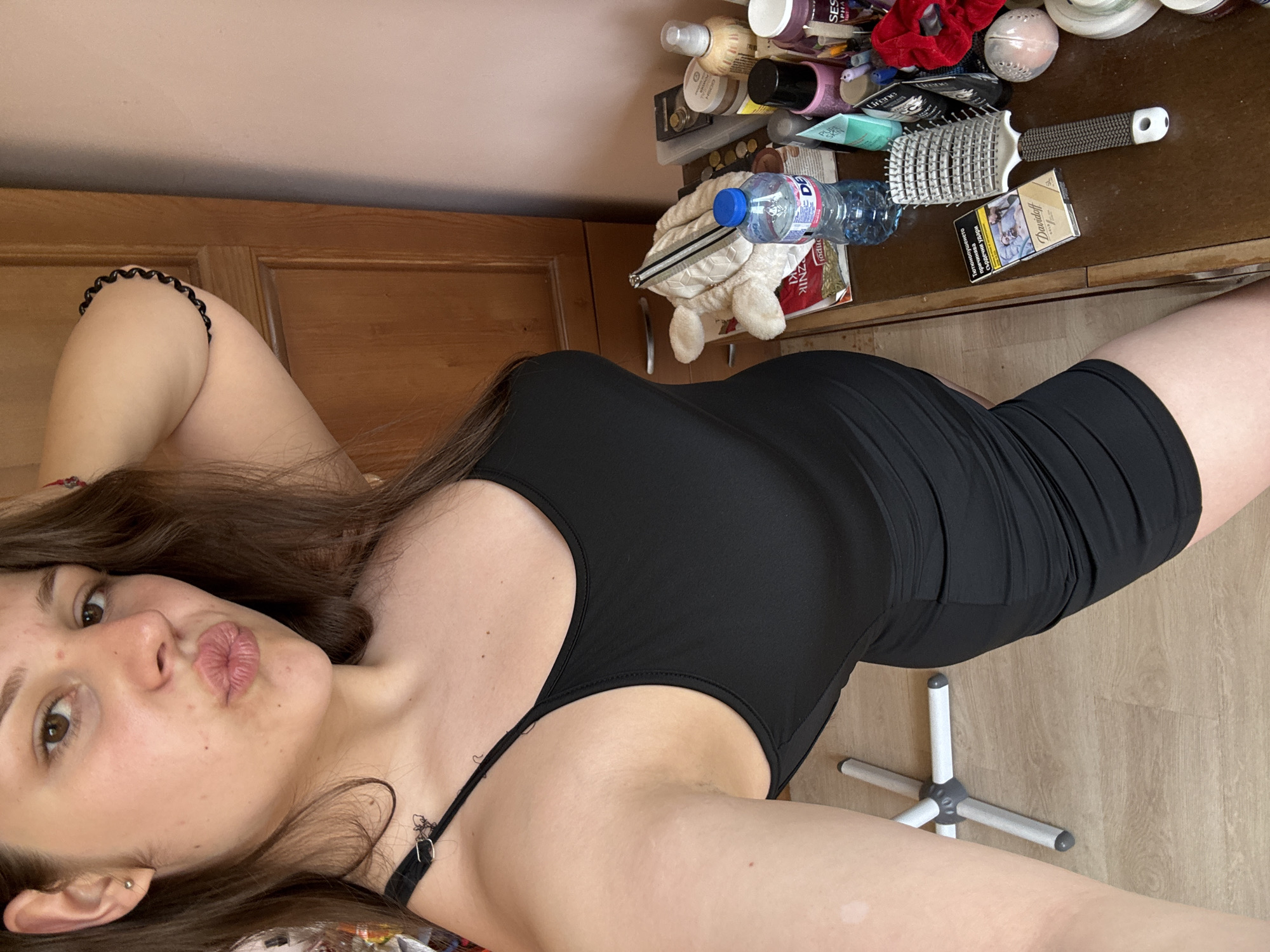 This media file is a selfie taken by a young woman in Prodanovtsi, Bulgaria. She is lying down, with her body positioned diagonally across the frame. She is wearing a black, form-fitting dress. Her hair is long and brown, falling around her shoulders. The woman is puckering her lips towards the camera, with one hand resting on her head.
In the background, there is a wooden dresser or table covered in various items. These include a hairbrush, a bottle of water, and several cosmetic products. A pack of cigarettes, specifically "Davidoff" brand, is also visible. The room appears to be lit by natural light, suggesting it is daytime. The floor is made of wooden planks. The overall mood is casual and relaxed.