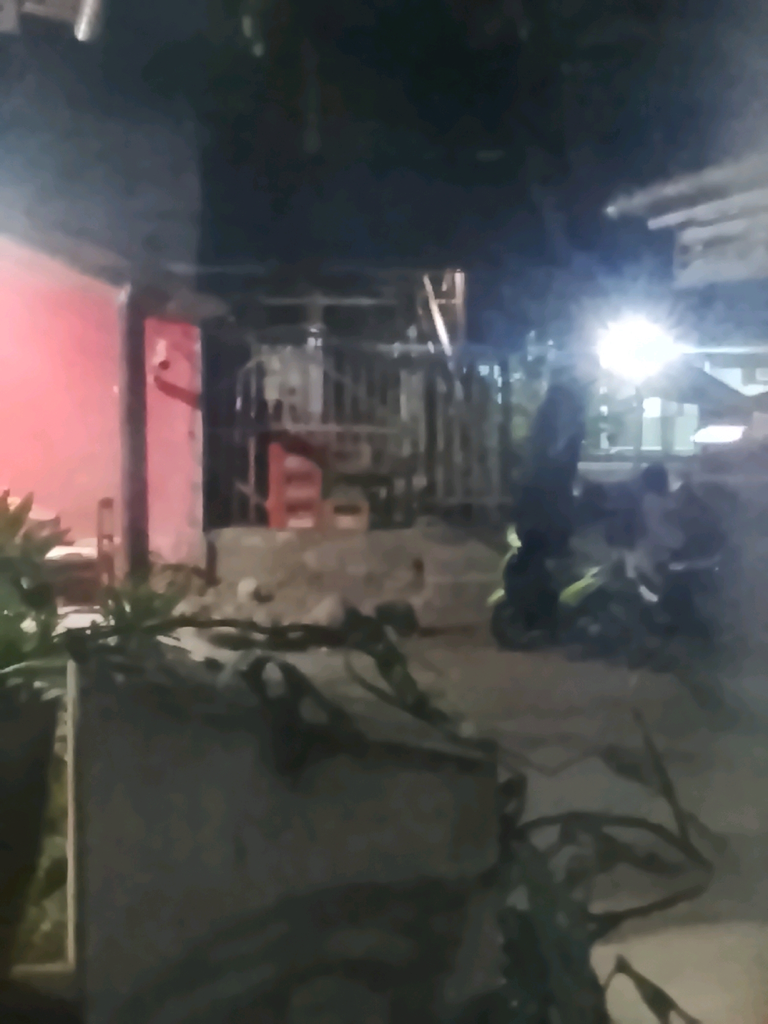 The image depicts an outdoor scene at night, likely in Ujungrusi, Indonesia, with a strong emphasis on artificial lighting that creates high contrast and obscures details. In the foreground, a dark, irregularly shaped object covered in what appears to be dark fabric or netting dominates the lower portion of the frame. To the left, a red wall with a vertical pipe is visible, partially obscured by greenery.
The middle ground shows a concrete structure with debris scattered on it, suggesting some disarray. Behind this, a metal fence or gate separates the area. A motorcycle, predominantly green, is parked on the ground, with a person dressed in dark clothing standing next to it, facing away from the camera. Another motorcycle is partially visible to the right.
The background is heavily influenced by bright artificial lights, including a particularly intense light source on the right, which creates lens flare and washes out much of the detail in that area. Structures are faintly discernible behind the fence, including what appears to be a building with a white exterior. The overall impression is one of a dimly lit, potentially utilitarian or residential area at night.