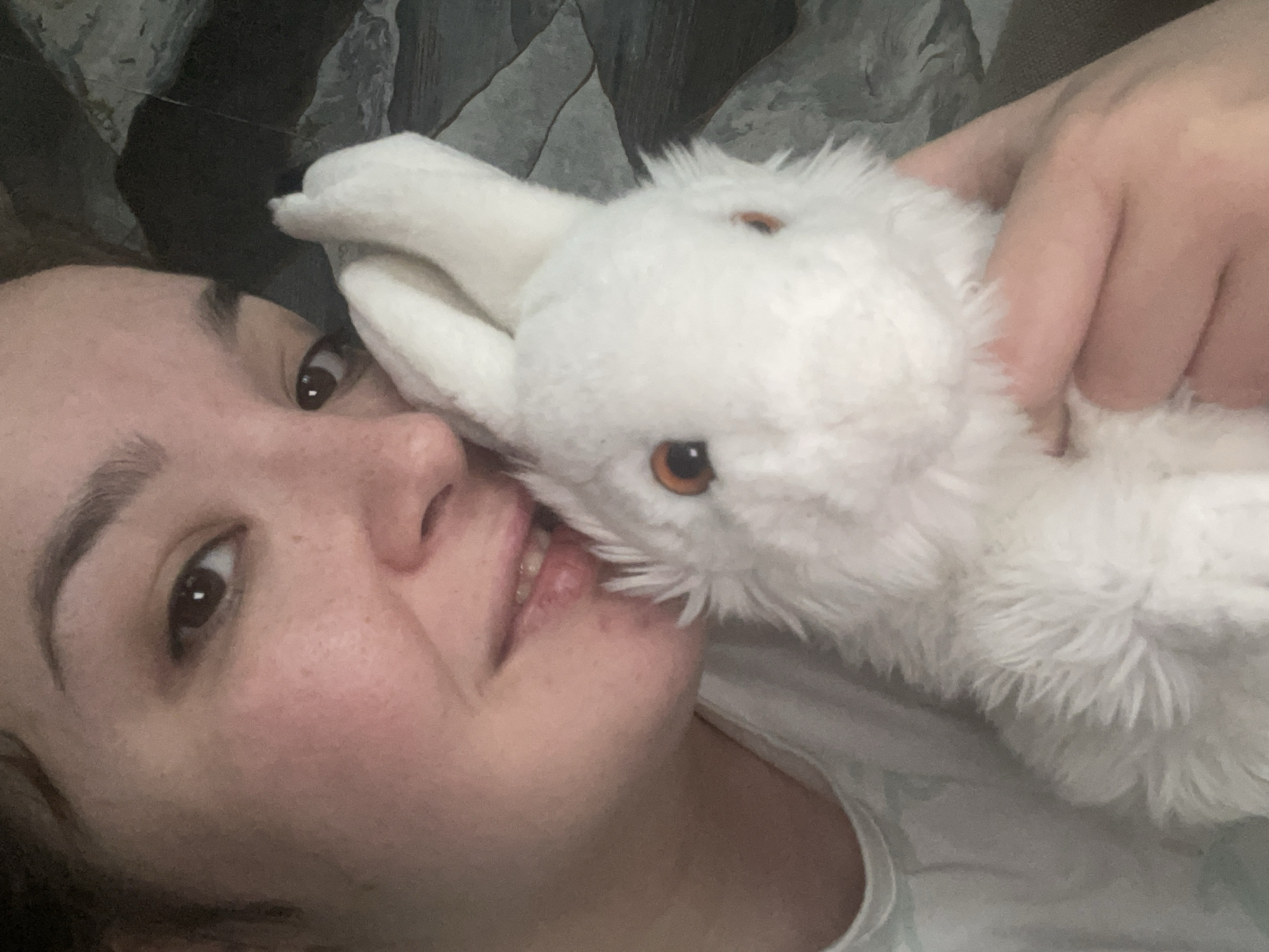 A close-up, portrait-style shot features a person with light skin and dark hair, lying down and holding a white, plush rabbit toy. The person's face is turned towards the camera, with their eyes open and lips slightly parted. The rabbit toy is positioned against the person's face, held by a hand that is also visible. The background is a textured surface, possibly a blanket or wall. The setting is unknown.