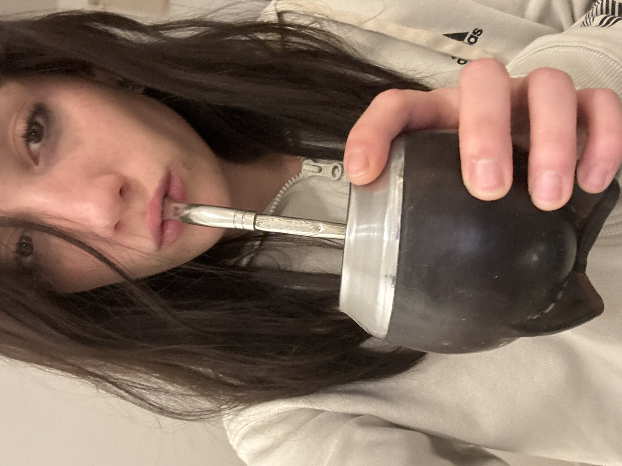A woman is holding a mate (a traditional South American caffeinated beverage) to her mouth, presumably to drink it. The mate is a black gourd with a metal rim and a metal straw (bombilla). The woman has dark hair and is wearing a light-colored sweatshirt, showing an Adidas logo. The photo is taken from a low angle. The location is unclear, but the mate indicates a connection to South America, especially Argentina, where it is a popular beverage. It's impossible to tell where in Buenos Aires this photo was taken, as there are no contextual clues beyond the mate.