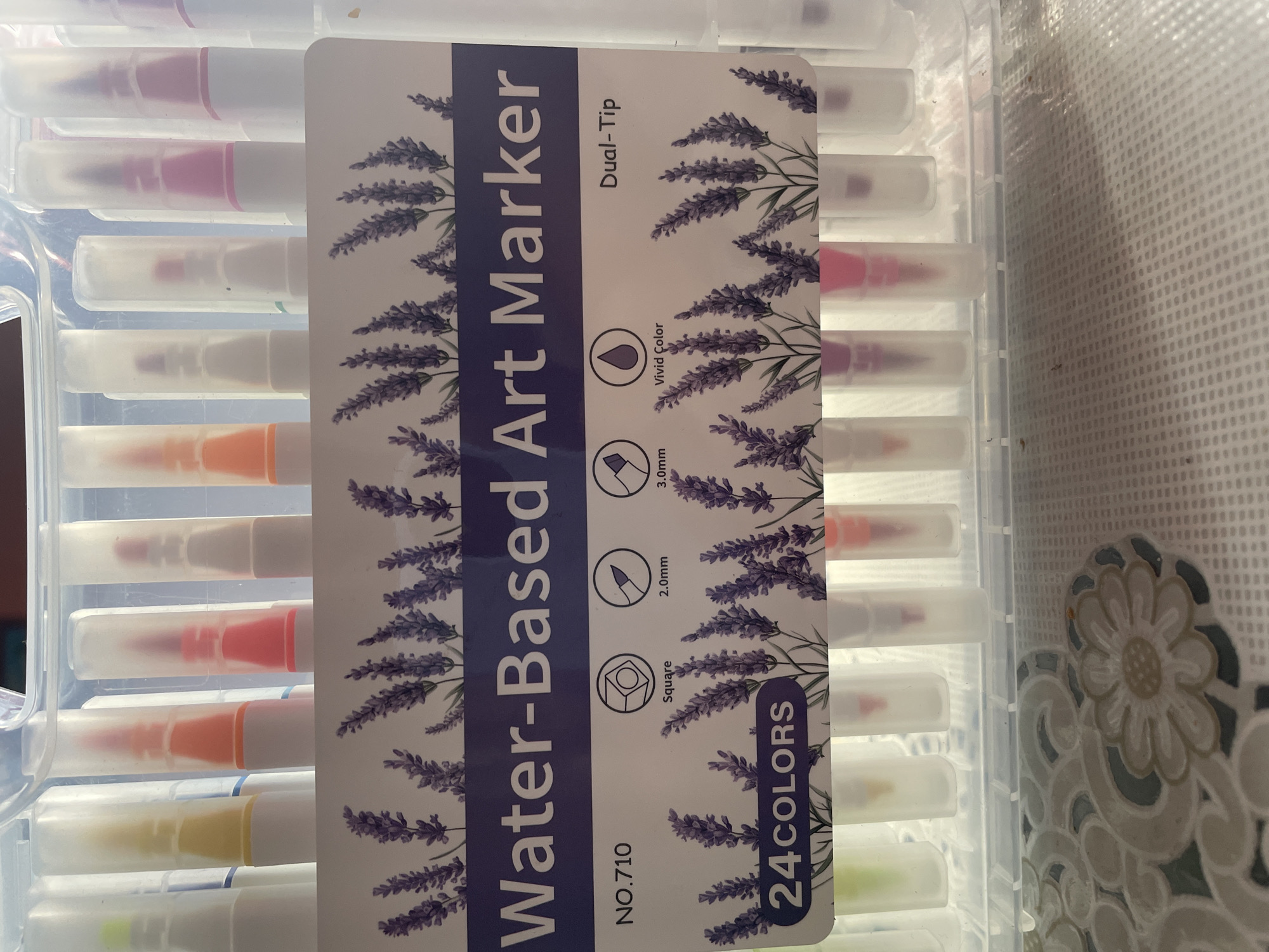 The image shows a clear plastic case containing a set of 24 dual-tip water-based art markers. The markers are arranged in two rows, with their caps on. The front of the case features a label with a lavender design. Text on the label reads "Water-Based Art Marker," "NO.710," "24COLORS," and "Dual-Tip." Below this text, there are icons indicating "Square," "2.0mm," "3.0mm," and "Vivid Color." The markers themselves have translucent white barrels, with colored caps and tips that are partially visible through the translucent outer casing. The background is blurred, but appears to be an indoor setting. There are no people or animals visible in the image. The location of Shpykolosy, Ukraine is not evident from the image.