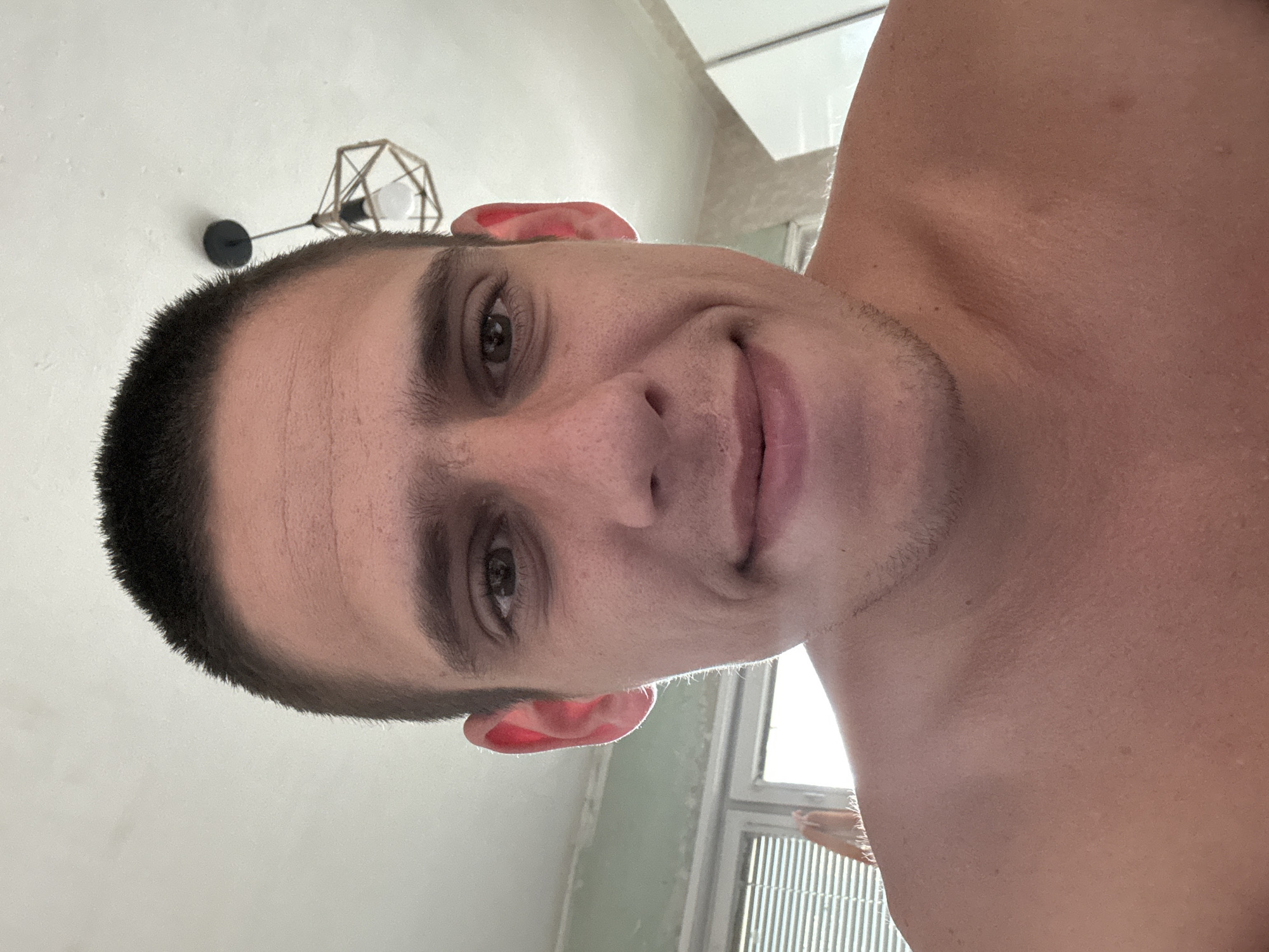 A close-up, head-and-shoulders selfie of a fair-skinned man with a buzzed haircut and light stubble on his chin and neck. He is looking directly at the camera with a slight smile. His eyes are open, and his eyebrows are visible. The man's ears are prominent, and his skin appears to have some minor blemishes and texture. He is shirtless, revealing his shoulders and upper chest.
The background suggests an indoor domestic setting. To the left, a window with blinds is visible, letting in bright natural light. Above the man's head, a light fixture with a geometric design hangs from the ceiling. The walls appear to be painted a neutral color, with some scuff marks or unevenness visible on the upper left wall. The overall lighting is bright, likely from the window and overhead light.
The image is taken from a low angle, looking slightly up at the subject. There are no other people or animals present in the frame. The composition is tight, focusing heavily on the man's face and upper body. No specific geographic markers or clues about the location in Sofia, Bulgaria, are discernible from the image itself.