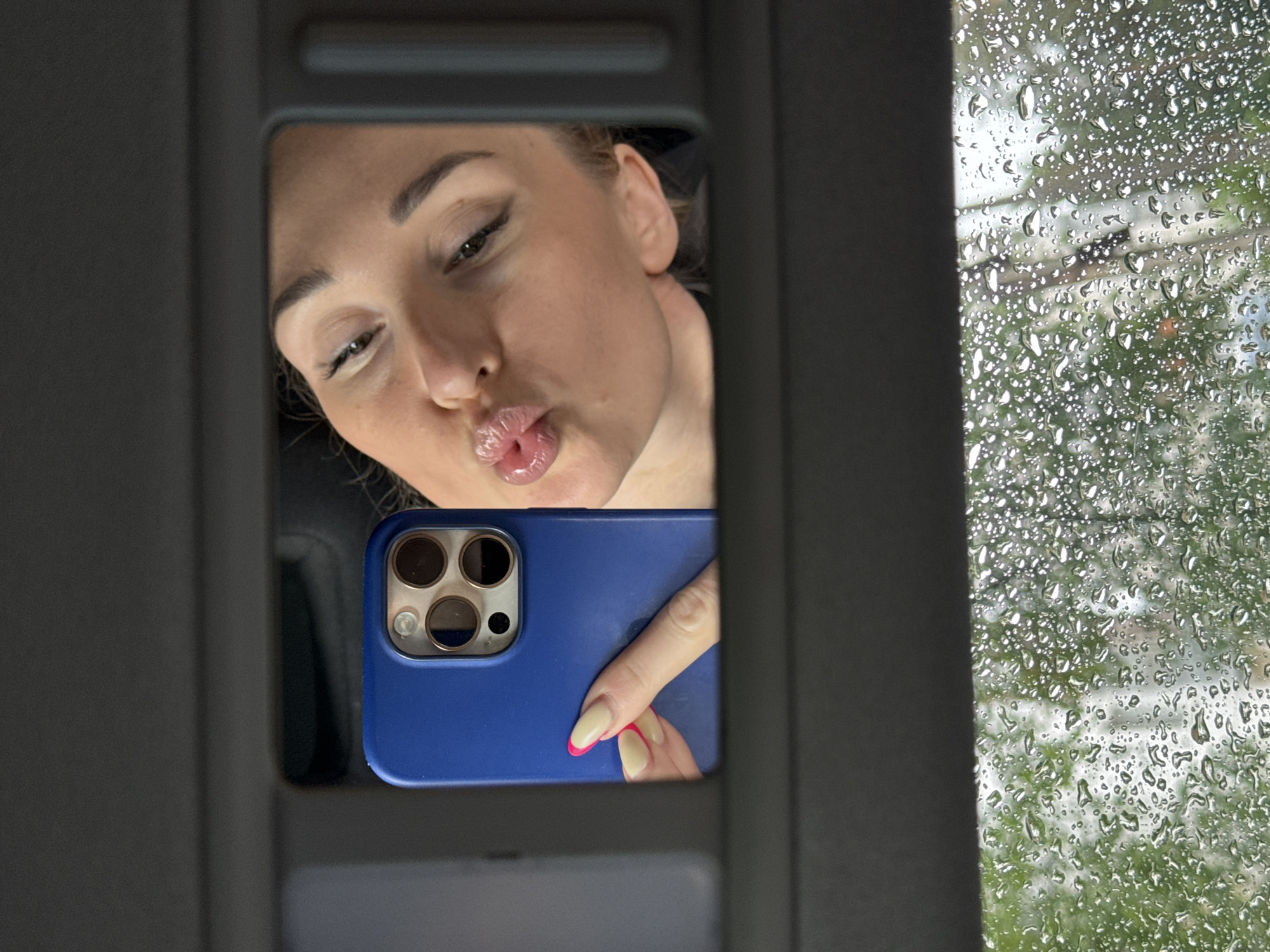The image displays a scene from inside a vehicle, likely in Chaikovsky, Russia, characterized by two distinct vertical halves. The left half shows a reflection in what appears to be a car's vanity mirror. In this reflection, a woman is captured in a selfie pose, looking down towards a smartphone she is holding. Her lips are pursed in a "kissing face" or "duck face" expression. She holds a smartphone encased in a prominent blue protective cover. Her visible hand reveals a manicure with beige-colored nails tipped in bright pink. The right half of the image features a window, heavily covered in numerous raindrops, indicating active rain or recent precipitation. Through the blurry water droplets, green foliage and an indistinct background are visible, suggesting trees or other natural elements outside the vehicle. The lighting suggests it is daytime. No visible text is present in the image.