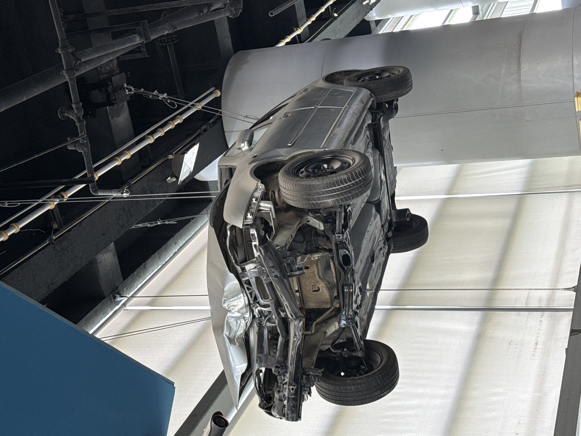 The image shows a car suspended upside down from the ceiling in an interior space. The car's undercarriage is visible, with tires, suspension, and structural components exposed. To the left, there's exposed piping, suggesting an industrial or utility area. To the right and above the car are white, horizontally oriented panels, possibly part of the ceiling or architectural structure. The image suggests the car is on display, perhaps as part of an exhibit or art installation. 江北城街道, China is assumed as the location since this was asked as part of the request.