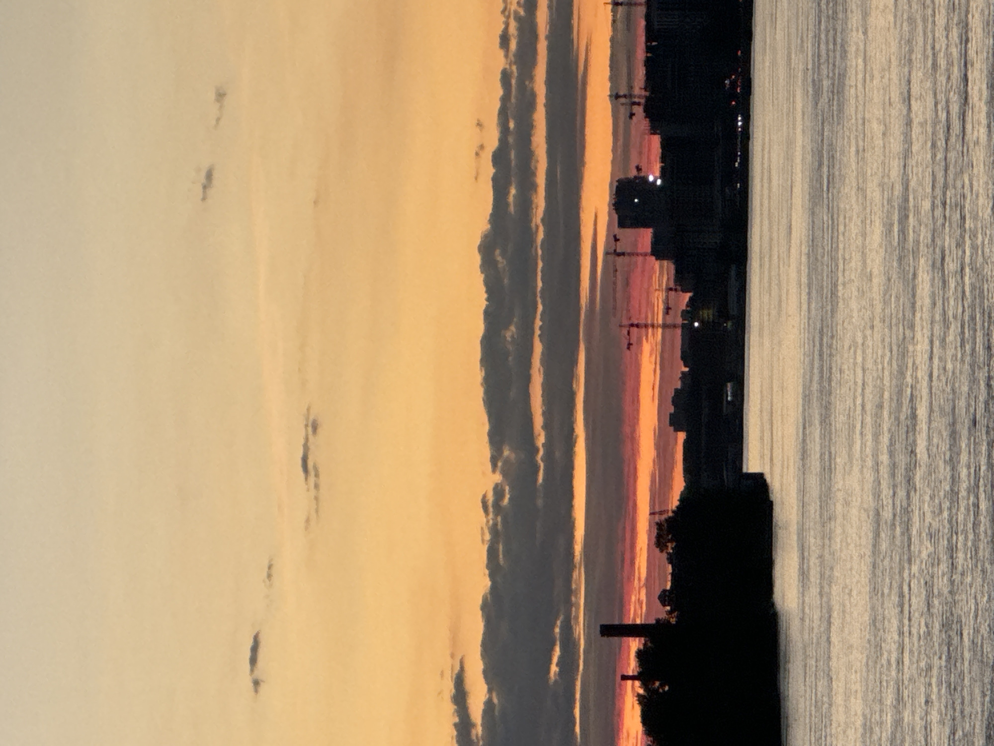 This is a photograph taken at dusk in Imeni Morozova, Russia, featuring a wide river in the foreground and a silhouetted cityscape on the horizon. The sky is painted with a gradient of warm colors, transitioning from a pale orange at the top to deeper reds and pinks near the horizon, with dark gray clouds scattered across the sky.
On the left side of the cityscape, a large, dark industrial chimney stands prominently against the sky, accompanied by silhouetted trees and buildings. Further to the right, the skyline is dominated by the silhouettes of numerous construction cranes and various buildings, suggesting ongoing development. The river in the foreground reflects the colors of the sky and appears calm with gentle ripples. The lighting conditions indicate it is either sunrise or sunset, with the sun below the horizon casting a warm glow. There are no people or discernible actions visible in the image.