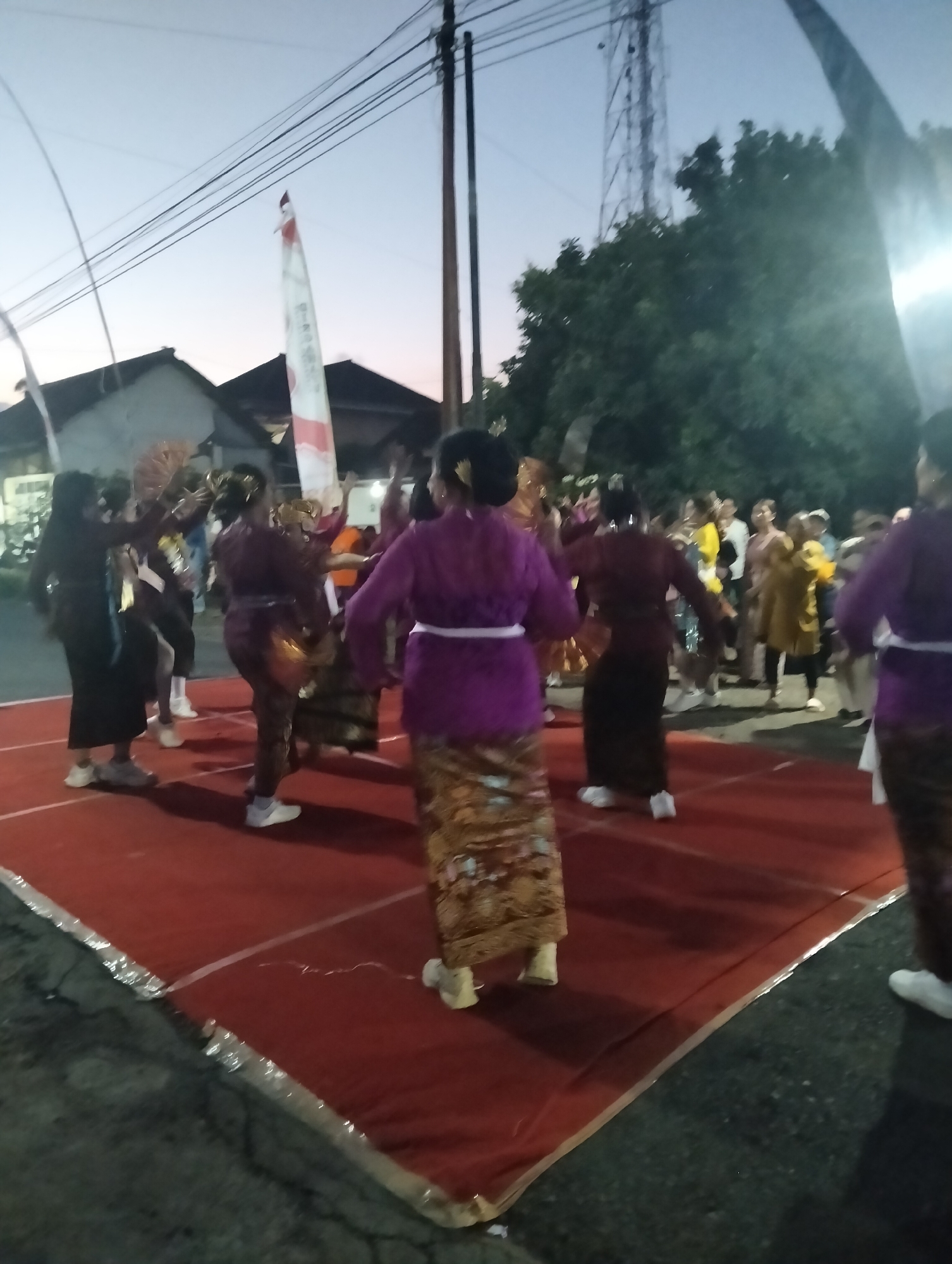 A group of people, primarily women, are engaged in a traditional dance performance on a raised red carpet stage. The dancers are dressed in colorful, traditional attire, including patterned skirts and blouses. Some dancers are holding fans, adding to the visual spectacle. In the background, there are houses, trees, and utility poles, suggesting an outdoor setting in a residential area. A flag, possibly of Indonesia given the red and white colors, is visible on a pole. The sky appears to be twilight, with a soft, diffused light illuminating the scene. The overall impression is of a cultural event or celebration taking place in the town of Sumbersewu, Indonesia.
