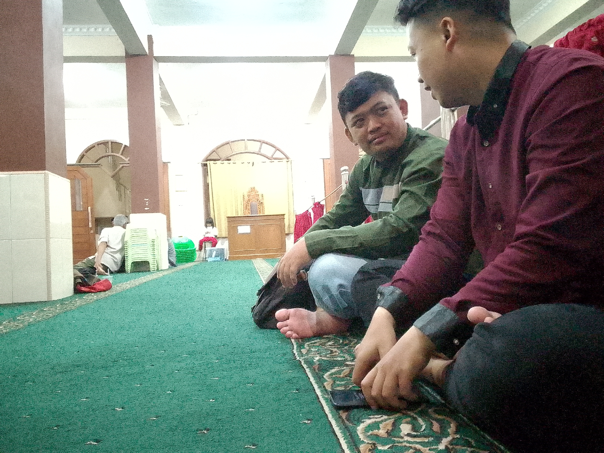 Two young men are seated on a green carpet inside a building that appears to be a mosque in Kertajaya, Indonesia. The man on the left is wearing a green long-sleeved shirt and blue jeans, and he is looking towards the right. The man on the right is wearing a burgundy long-sleeved shirt and black pants, and he is also looking to the right. In the background, there are pillars, arched doorways, and several stacks of green plastic chairs. Other people are visible in the background, including one person kneeling and another person sitting. The overall lighting suggests it is daytime.