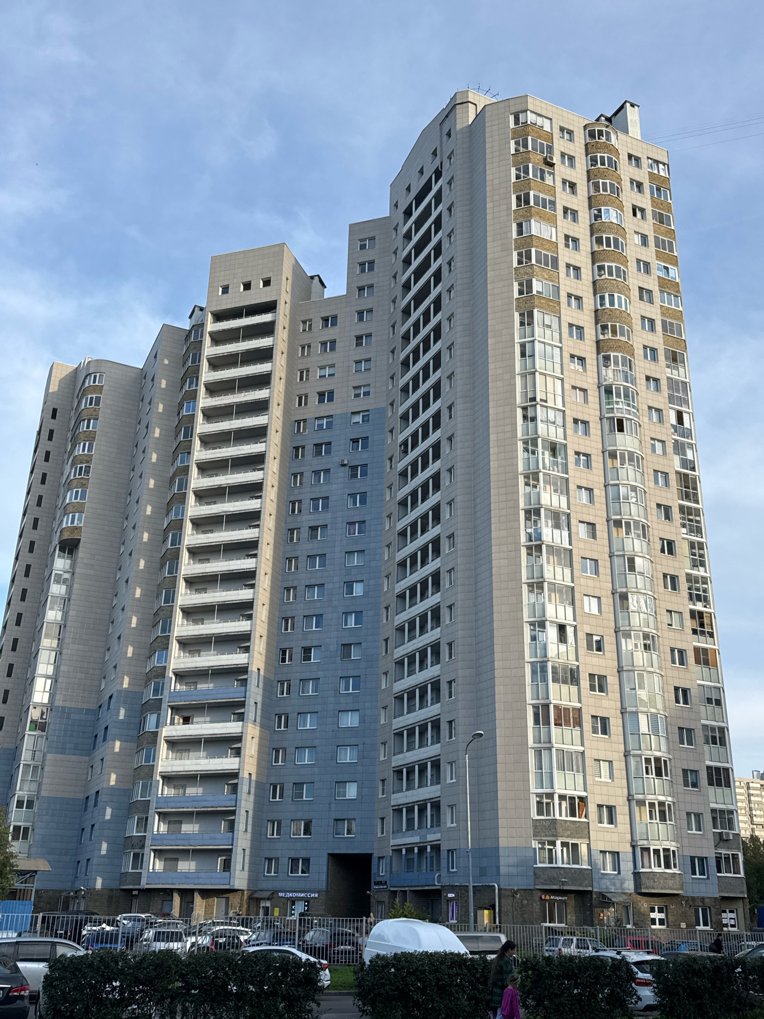 An exterior, eye-level, medium shot captures a modern, multi-story apartment building against a sky with scattered clouds. The building is composed of several sections with varying heights and architectural styles. The main section on the right is tall and rectangular, clad in light gray and beige panels, with a facade featuring numerous windows and several curved balcony areas. The left section of the building is also tall but appears to have a more segmented design, with repeating vertical rows of balconies and windows. This section is primarily clad in light gray panels with blue accents. The lower levels of the building appear to have commercial spaces with signage. In the foreground, a row of dark green bushes separates the building from a parking lot filled with various cars, including sedans, SUVs, and a white van. A few people are visible near the bushes and in the parking lot. The overall scene suggests a residential urban environment.
