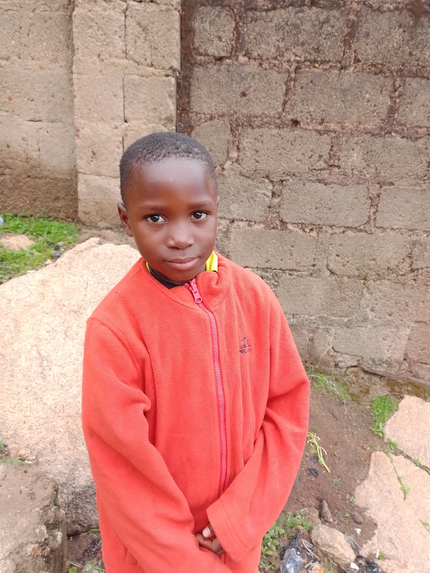 A young boy with dark skin and short black hair stands in front of a brick wall and a large rock. He is wearing an orange fleece jacket with a zipper. The boy is looking directly at the camera with a neutral expression. The background is slightly out of focus, but appears to be an outdoor setting with some greenery and debris on the ground.