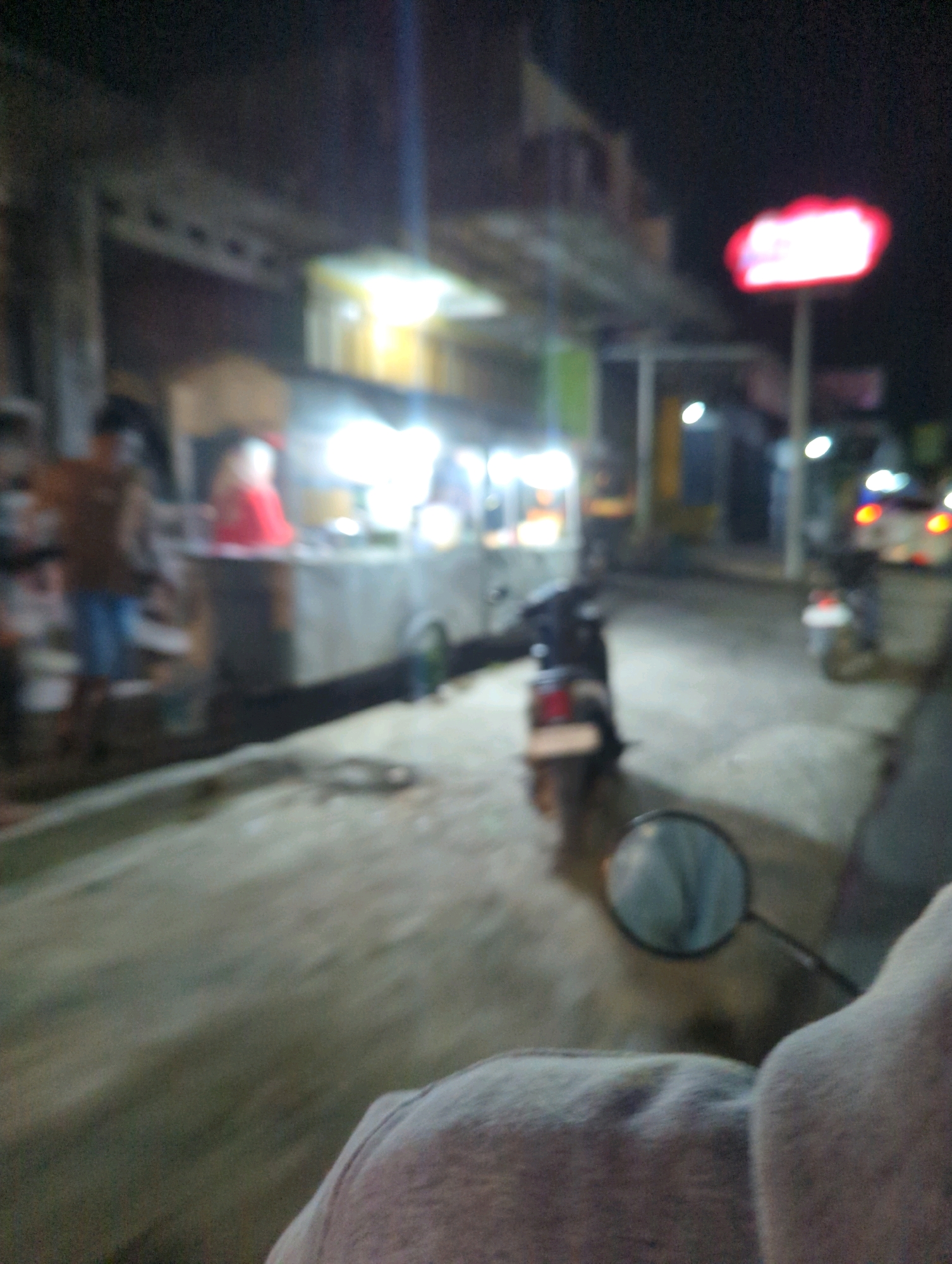 The image depicts a blurry night scene, possibly a street in Kacangan, Indonesia. In the foreground, a person's shoulder and upper arm are visible, suggesting the photo is taken from the perspective of someone riding a motorcycle or sitting on one. A motorcycle's handlebar and mirror are also in the foreground, with the mirror reflecting a hazy view of the surroundings. In the mid-ground, a street vendor's stall is illuminated by bright lights. Two figures are partially visible at the stall: one wearing a brown shirt and blue shorts, and another in a red garment. Several motorcycles are parked or present in the scene. In the background, there is a building structure and a prominent red sign with white lettering, possibly an advertisement. The overall impression is a lively, commercial night street scene.