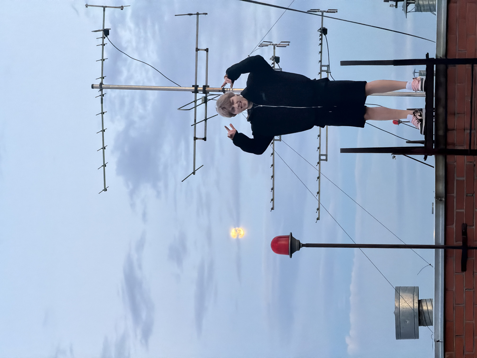 A young person with blonde hair, wearing a black zip-up hoodie, black shorts, and pink and black sneakers, stands on a metal structure on a rooftop. They are making a peace sign with each hand. Behind them, a large television antenna is mounted on a pole, with wires extending downwards. To the left of the person, a red beacon light is visible on a pole, and further to the left, a cylindrical metal vent protrudes from the rooftop. In the sky, there are scattered clouds and what appears to be the moon glowing faintly. The rooftop is constructed of red brick. The scene is set against a cloudy sky, likely in the city of Khimki, Russia.