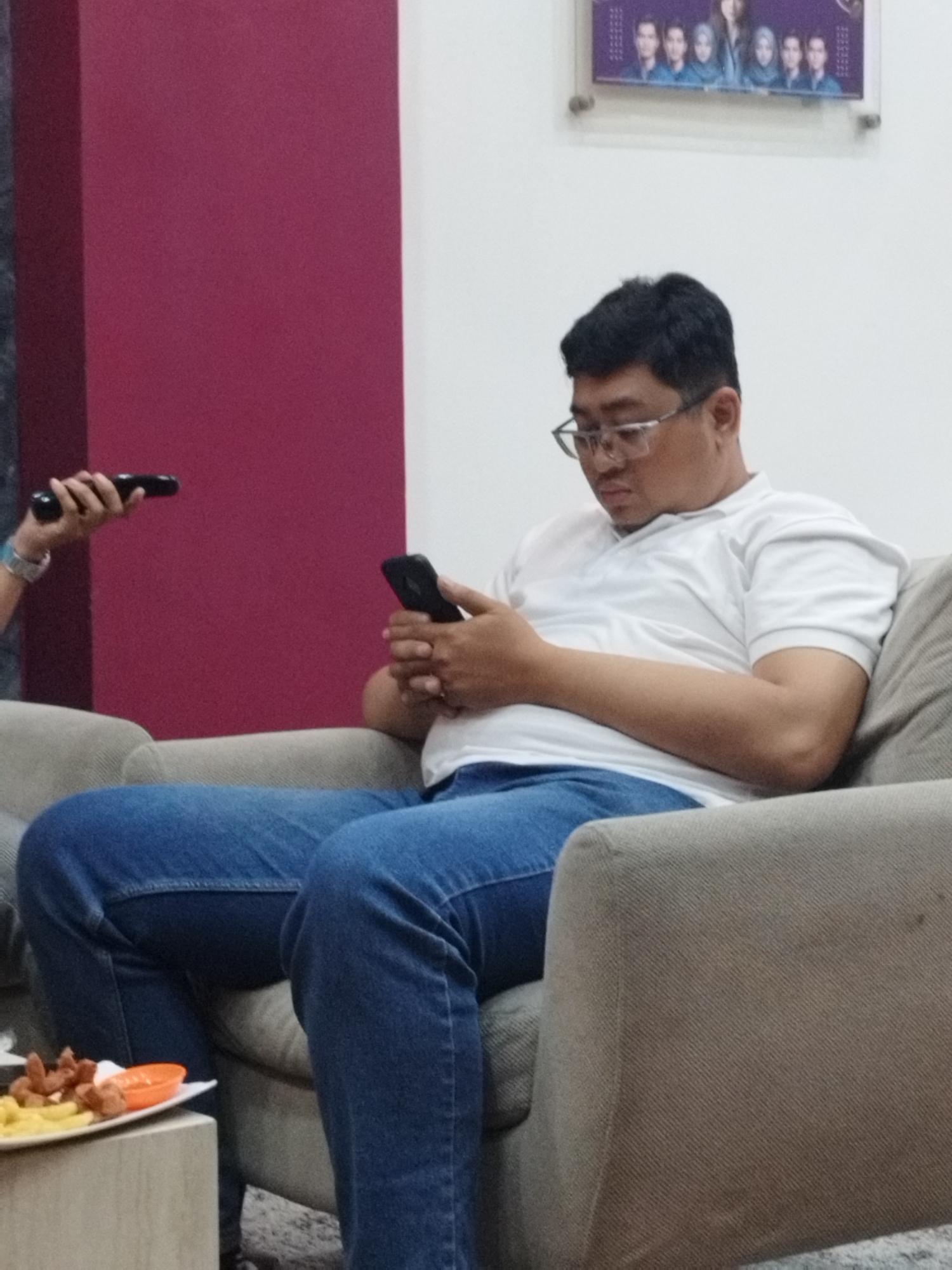 A man wearing glasses, a white polo shirt, and blue jeans is sitting on a sofa, looking at his smartphone. To his left, another person's hand is visible holding a black object, possibly another smartphone or remote control. In the foreground, a white plate holds food, including french fries and what appears to be sausage. Behind the man, a framed picture hangs on a light-colored wall, displaying a group of individuals. To the left of the frame, a tall, dark red rectangular structure is visible. The scene appears to be indoors, possibly in a cafe or lounge area in South Jakarta, Indonesia, given the context of the image's origin.