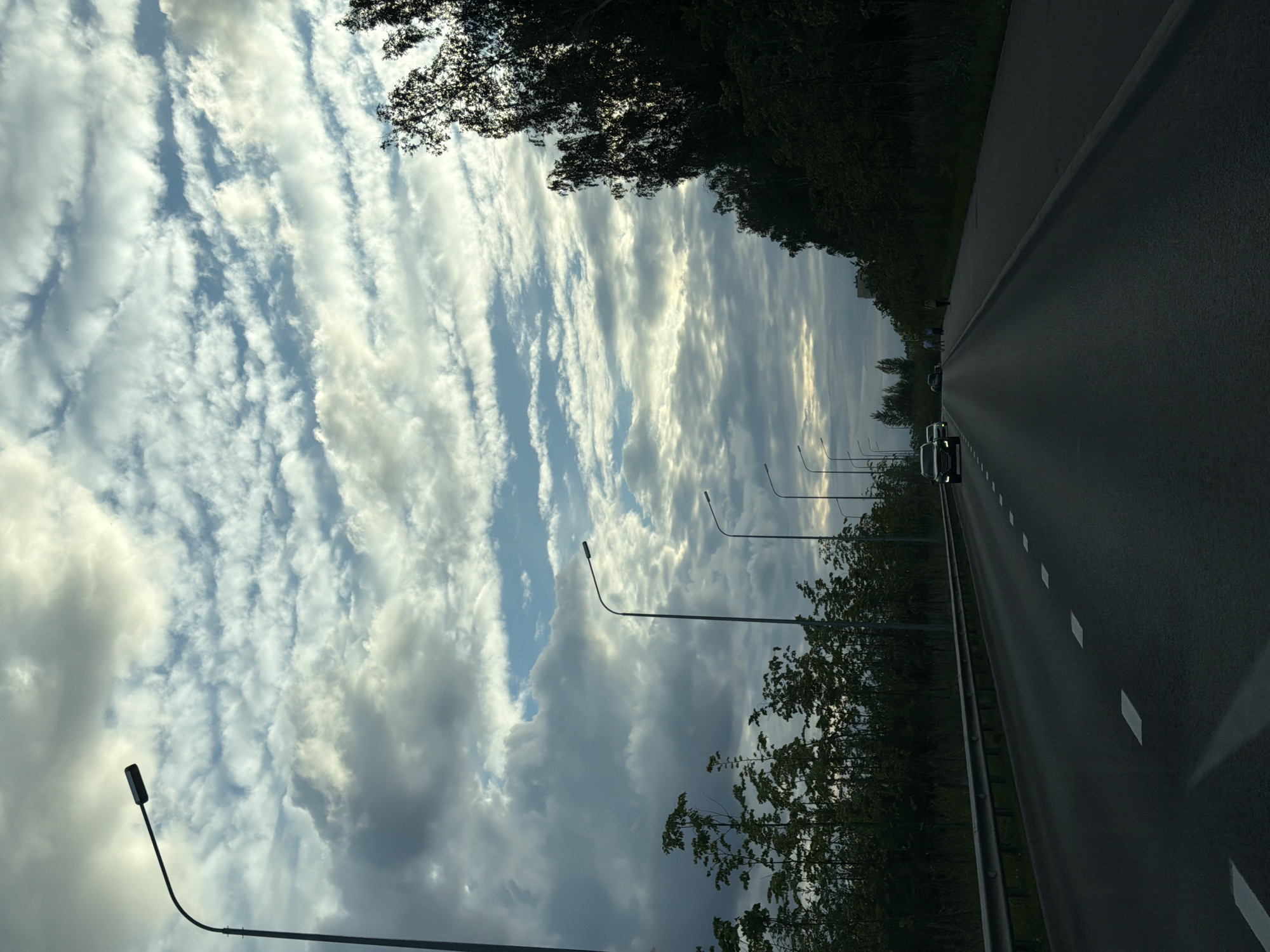 The image depicts a highway scene, likely within or near Kazan, Russia, under a dramatic, cloud-filled sky. The perspective is from a moving vehicle, looking forward along the road. Several streetlights line the left side of the highway, their tall poles stretching towards the sky. On the right, dense trees and foliage form a natural barrier along the roadside. The highway itself is paved, with white dashed lines indicating lanes. Two cars are visible in the distance, traveling in the same direction as the viewer. A few pedestrians are also seen on the right side of the road, near the tree line. The clouds are varied, with patches of blue sky peeking through, and some illuminated by an unseen light source, possibly the sun beginning to set or rise. The overall atmosphere suggests an urban or semi-urban setting on a transit route.