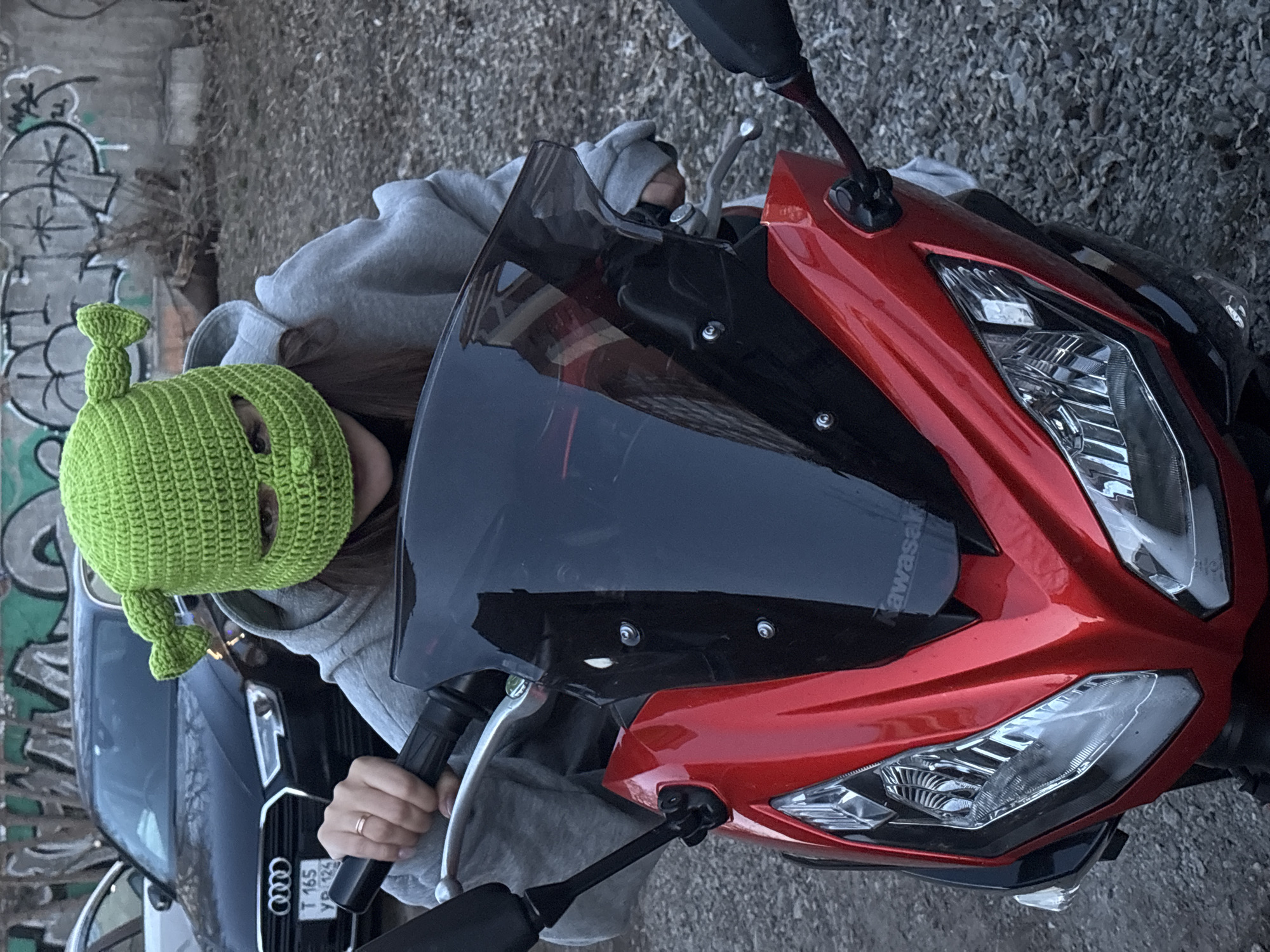 An outdoor, slightly overhead shot captures a person on a red and black Kawasaki sportbike in what appears to be an urban or semi-urban environment in Krasnoyarsk, Russia. The individual, whose gender is not explicitly discernible but appears to be female, is wearing a light grey hooded sweatshirt and a distinctive bright green knitted balaclava-style hat designed to resemble Shrek, complete with eye holes and ear-like protrusions. Only one eye is visible through the left eyehole of the hat. The person's left hand, adorned with a ring on the ring finger, is gripping the motorcycle's handlebar.
The motorcycle is a Kawasaki sportbike, identifiable by the "Kawasaki" text visible on its black upper fairing, contrasted with its vibrant red bodywork. It features a dark windscreen and prominent front headlights. In the background, a dark-colored Audi sedan is partially visible, displaying a Russian license plate with the characters "T 165 УР 124". Further background elements include a concrete wall marked with graffiti and sparse, dry vegetation. The scene is lit by natural daylight, indicating a daytime setting. The playful Shrek hat serves as a notable, lighthearted detail in the image.
