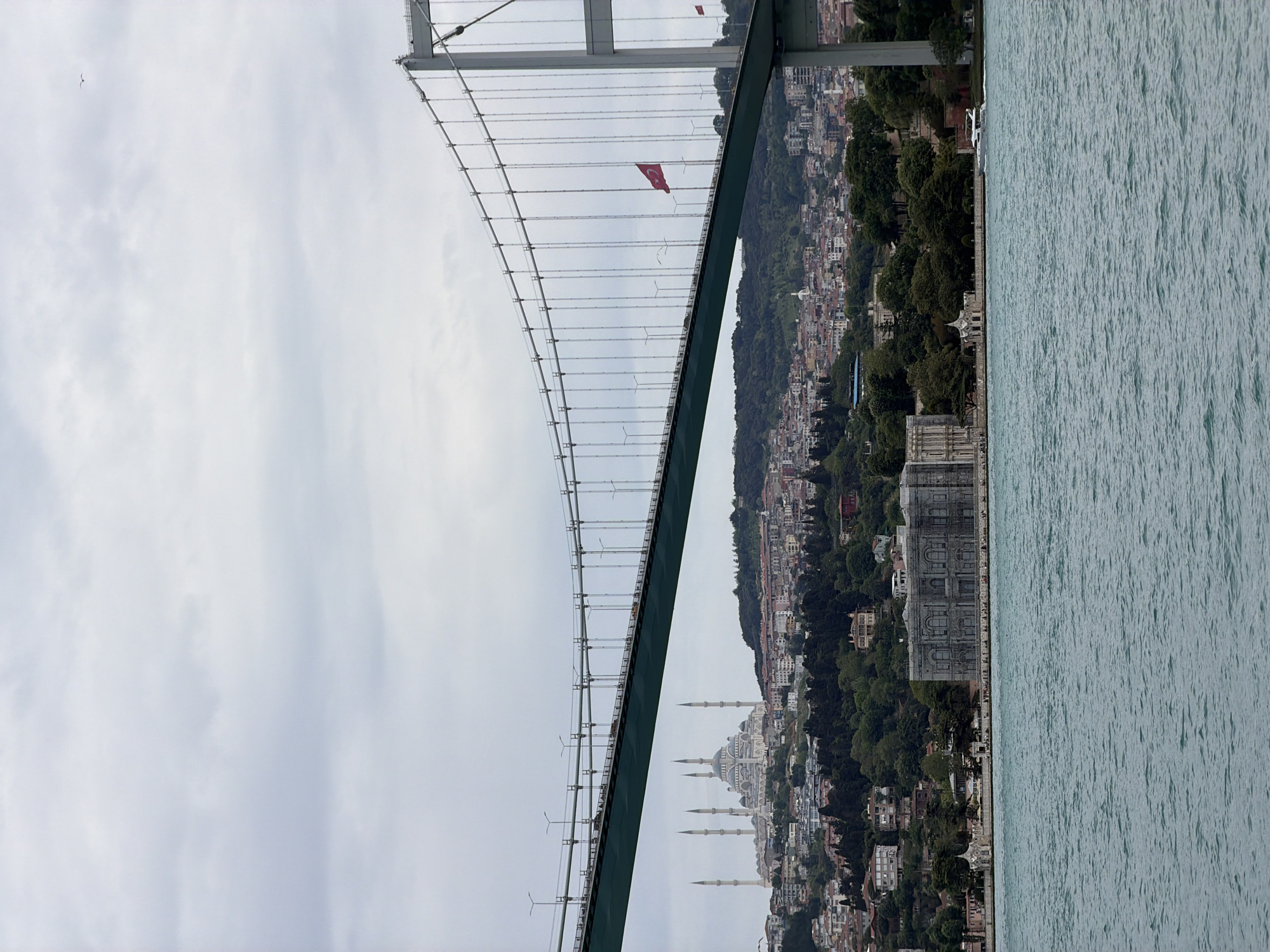 The photograph depicts a partial view of a bridge, likely the Bosphorus Bridge, in Istanbul, Turkey. The bridge structure is a suspension bridge with cables visible on the left. A Turkish flag is hanging from the bridge structure. On the right, the cityscape of Beşiktaş is visible, including densely packed buildings and trees. A large mosque with multiple minarets is also prominently visible. The foreground to the right is water. The sky is overcast. A bird is visible in the sky.