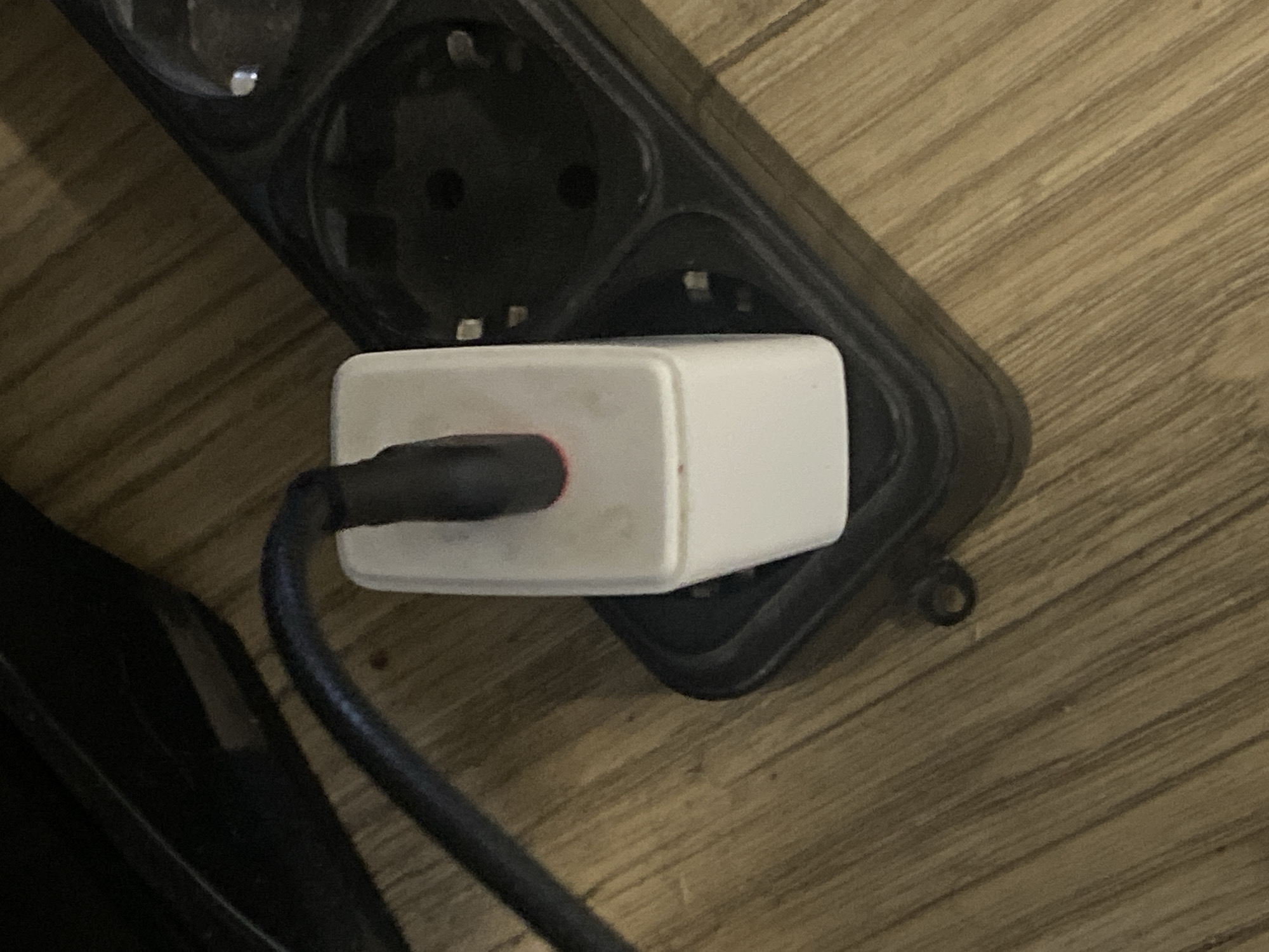 The image shows a white power adapter plugged into a black power strip. The power strip is on a wooden floor. The power adapter has a black cable connected to it. The power strip has multiple sockets, some of which are empty. The scene appears to be indoors, possibly in a home. The image does not provide any information about the city of Rozhny, Ukraine, or any people.