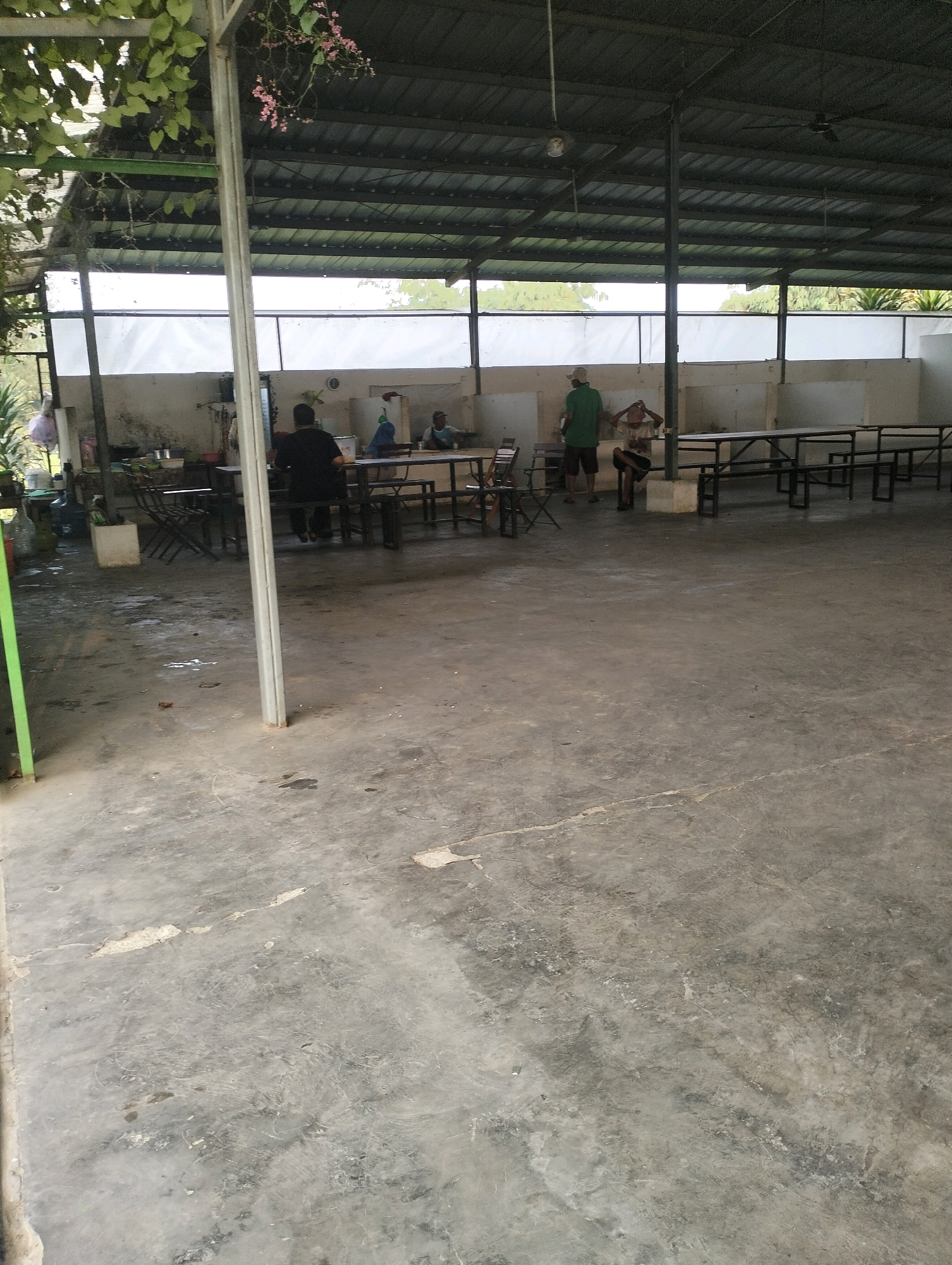 An outdoor, covered eating area in Gunung Sindur, Indonesia, features several tables and benches. In the background, three individuals are visible. One person, wearing a dark shirt, is seated at a table. Another, in a green shirt, stands near a table. The third person, wearing a light-colored shirt, is seated. The concrete floor is worn and stained. The roof is made of metal sheeting, supported by metal beams. Green foliage hangs from the ceiling on the left side of the image, and pink flowers are present. The area appears to be part of a larger establishment, possibly a restaurant or a community gathering space.