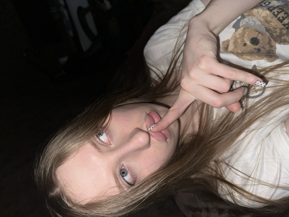 A young female subject with long, straight, light-brown hair and blue eyes is captured in a close-up portrait. She gazes upwards and to her left (viewer's right). Her lips are pursed in an 'O' shape, and the index finger of her right hand is lightly pressed against her upper lip, indicating a gesture of silence. Her fingernails are painted white with black polka dots. The dorsum of her right hand and wrist exhibits several faint, linear, reddish marks. She is wearing a light-colored short-sleeved t-shirt featuring a partial graphic print of a brown teddy bear and obscured text, including "KEEP G". The background is dark and out of focus, consistent with an indoor setting under direct frontal lighting. The image provides no visual data to confirm its specific location in Chaikovsky, Russia.