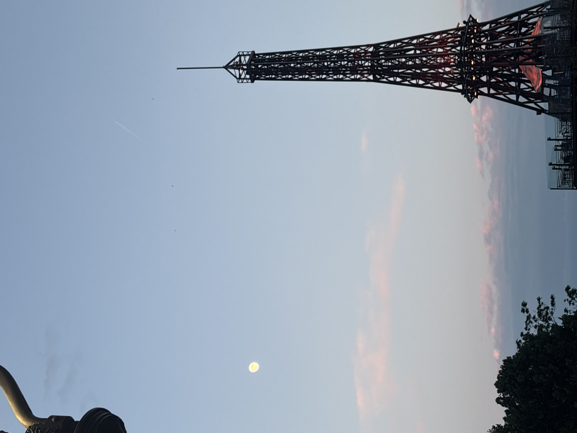 The image shows a partial view of what appears to be a replica of the Eiffel Tower against a twilight sky. The tower is constructed of dark metal and has some light illumination. Below the tower, there are some trees. To the left and below the tower, there is a dim moon. A contrail or airplane path is visible in the upper left portion of the sky. In the lower left is a statue or architectural feature. Based on the presence of the replica Eiffel Tower, it is possible that the location is Varna, Bulgaria, where such a replica exists.
