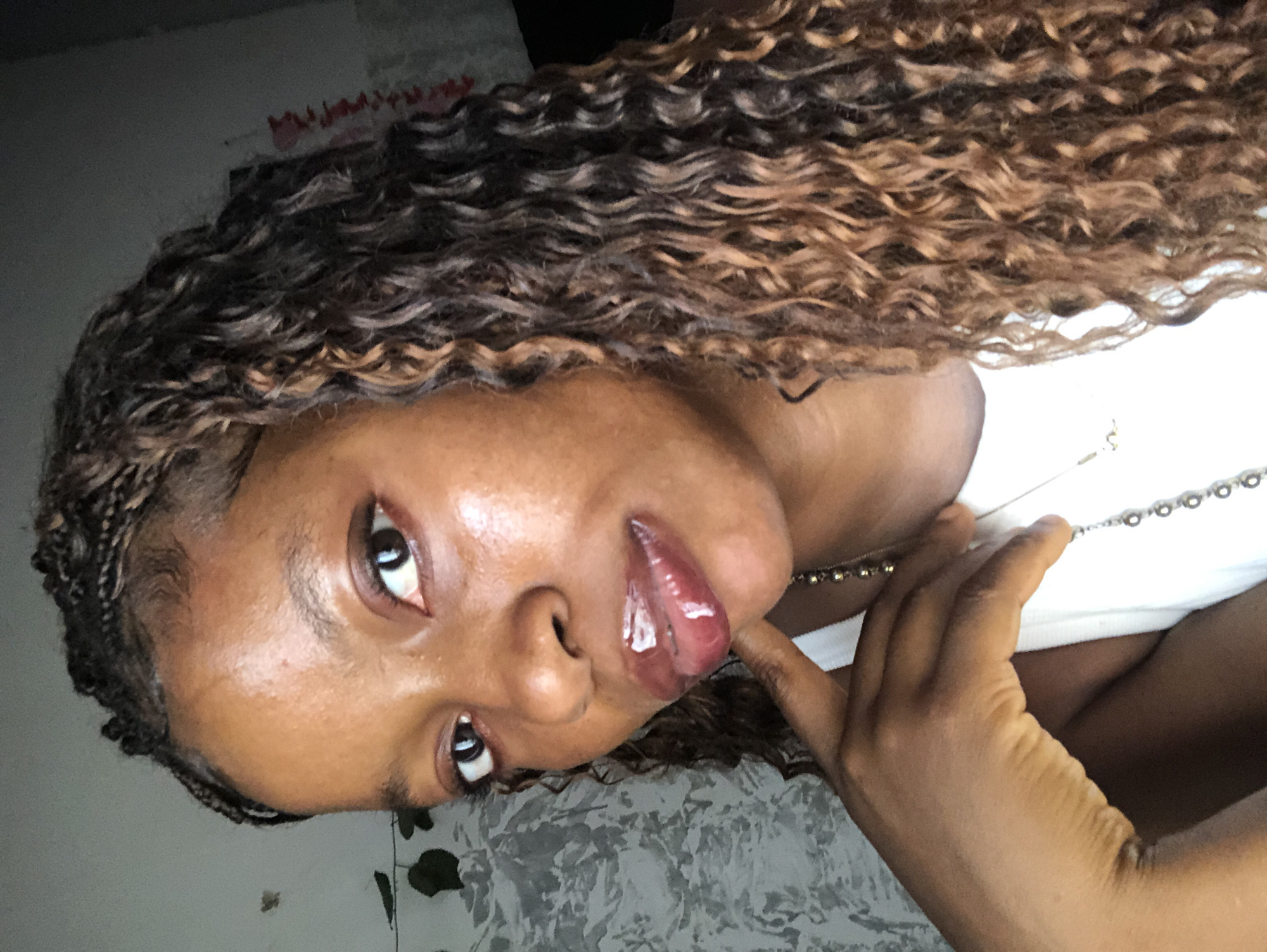 A woman with brown curly hair, braided at the forehead, is shown from the chest up. She is wearing a white tank top with a silver necklace. Her right hand is raised and touching her neck. Her face is slightly shiny. The location is Unknown, Nigeria. The background is a light gray wall with some indistinct decorations.