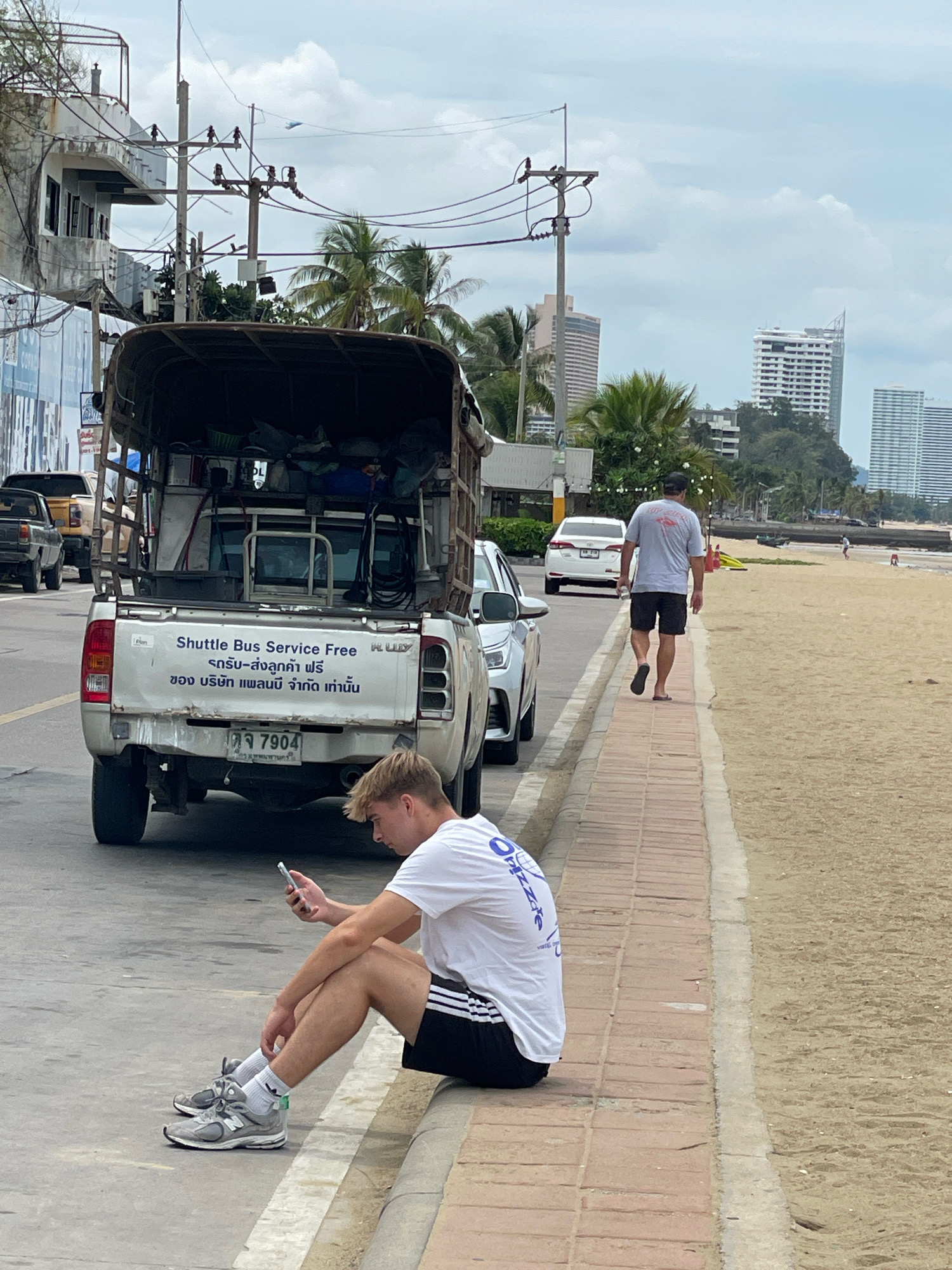 In Pattaya City, Thailand, a young man sits on a curb, looking at his phone, on a sidewalk next to a street. He is wearing a white t-shirt, black shorts with white stripes, white socks, and grey sneakers. A white pickup truck with a cage-like structure in the back is parked on the street to his left. The truck has text on its tailgate that reads "Shuttle Bus Service Free" in English and Thai. A man in a grey t-shirt and black shorts walks away from the camera on the sidewalk, towards the beach. The beach is to the right of the sidewalk, with sand and a few people in the distance. In the background, there are palm trees and tall buildings. The sky is overcast.