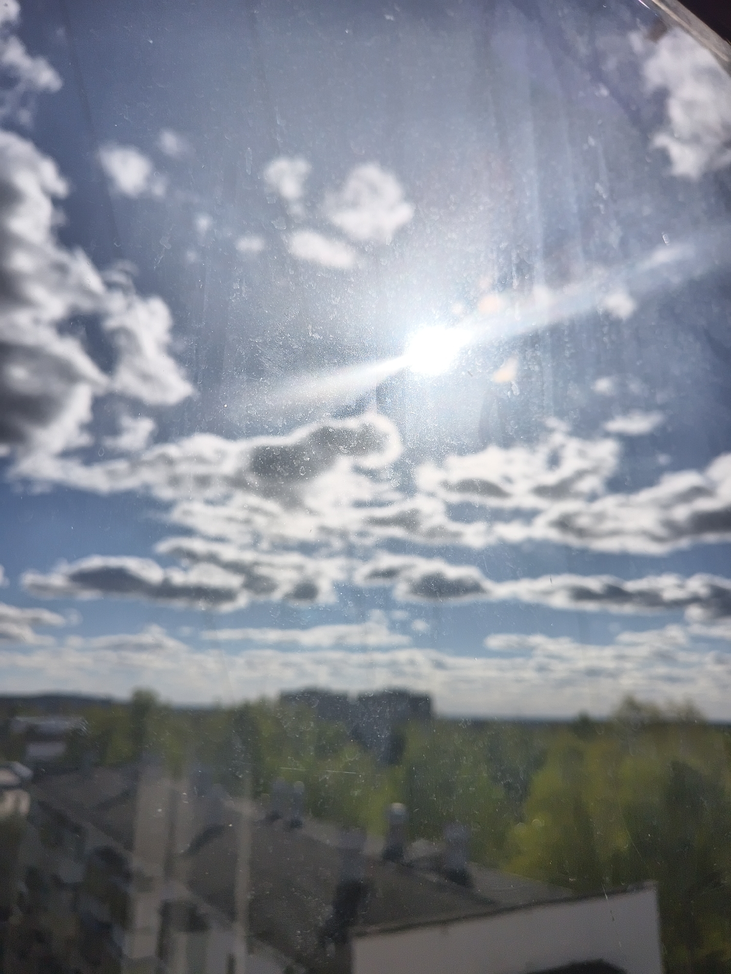 The image presents an outdoor scene viewed through a dirty window. The sky is visible, featuring scattered clouds and bright sunlight. Below the clouds, a distant cityscape is partially visible, consisting of buildings and trees. The foreground shows rooftops of buildings, likely part of the same urban area. The image has a blurred quality due to the dirt and imperfections on the windowpane. The location appears to be a city, but its specific identity is unknown.