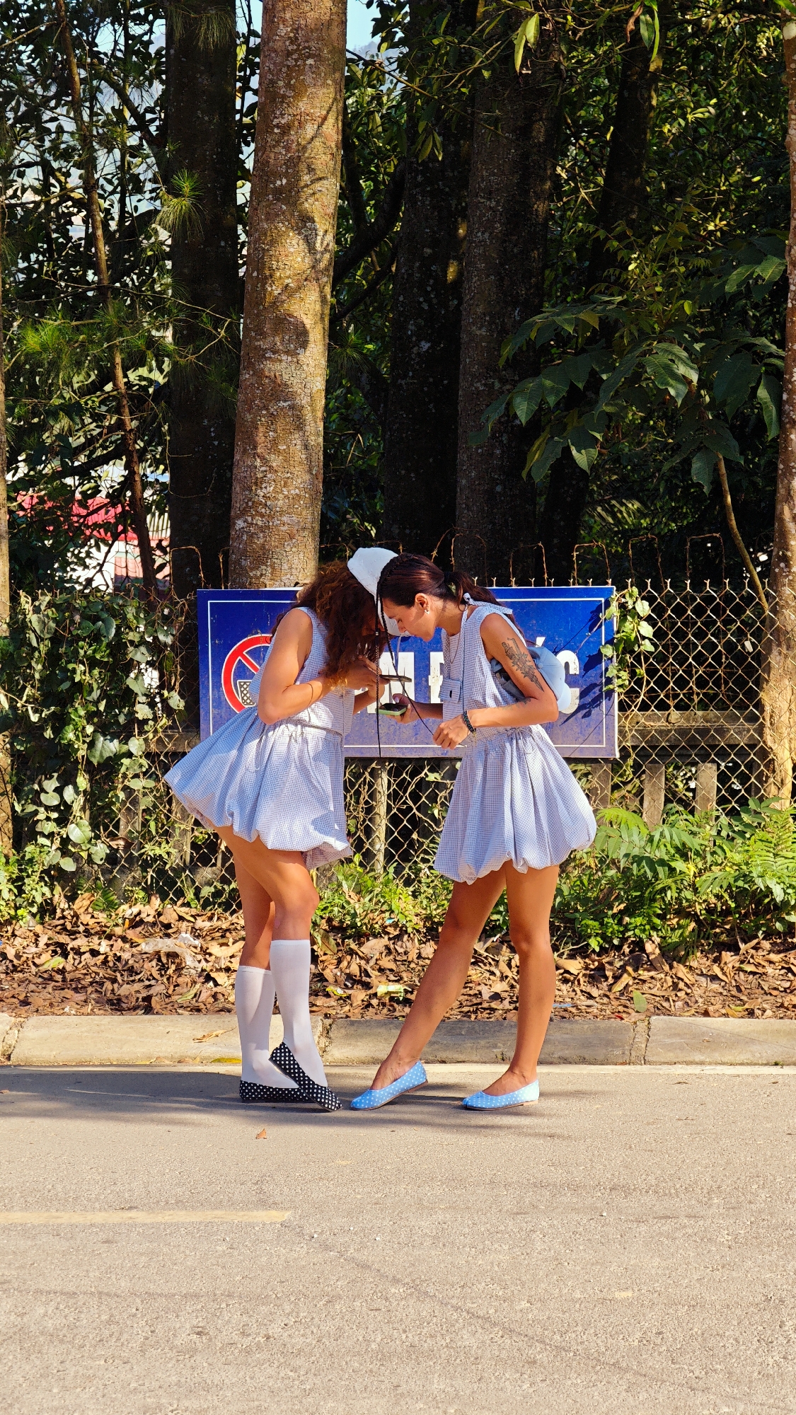 Two women stand on a paved road in front of a chain-link fence, trees, and foliage in Bắc Hà, Vietnam. They both wear light blue and white gingham dresses and white nurse caps. The woman on the left wears white knee-high socks and black and white polka-dot flats; the woman on the right wears light blue flats. They are both looking at a small object held between them. Behind them is a blue sign with a red circle and slash over a symbol, possibly indicating a restricted activity. The environment includes trees, foliage, and a fence.