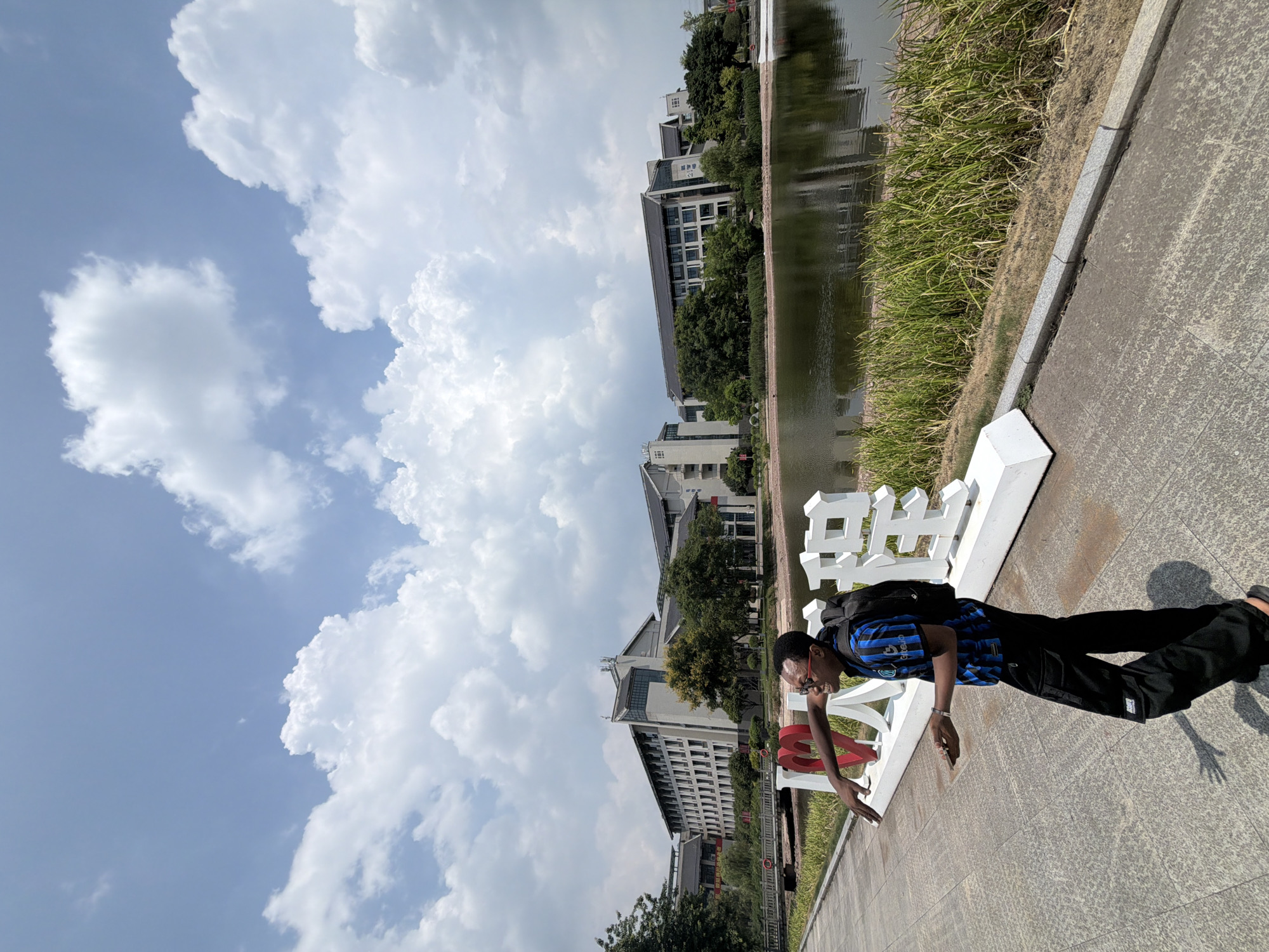 This image depicts a young Black man standing on a paved walkway next to a body of water, with buildings and trees in the background. The man is wearing a blue and black striped shirt, black cargo pants, and a black backpack. He is wearing glasses and has his right arm extended, with his hand open. In front of him, there are large white three-dimensional characters and a red heart shape, forming a decorative sign. The environment appears to be a park or campus, with lush green grass and reeds along the water's edge. The sky is blue with prominent white clouds. The buildings in the background are modern in design, with multiple stories and dark-colored roofs. The water reflects the sky and the surrounding structures. The overall scene suggests a bright, sunny day in an outdoor setting. The sign with the characters and heart likely reads "I love Jiangjin" (我爱江津), indicating the location as Jiangjin, China.