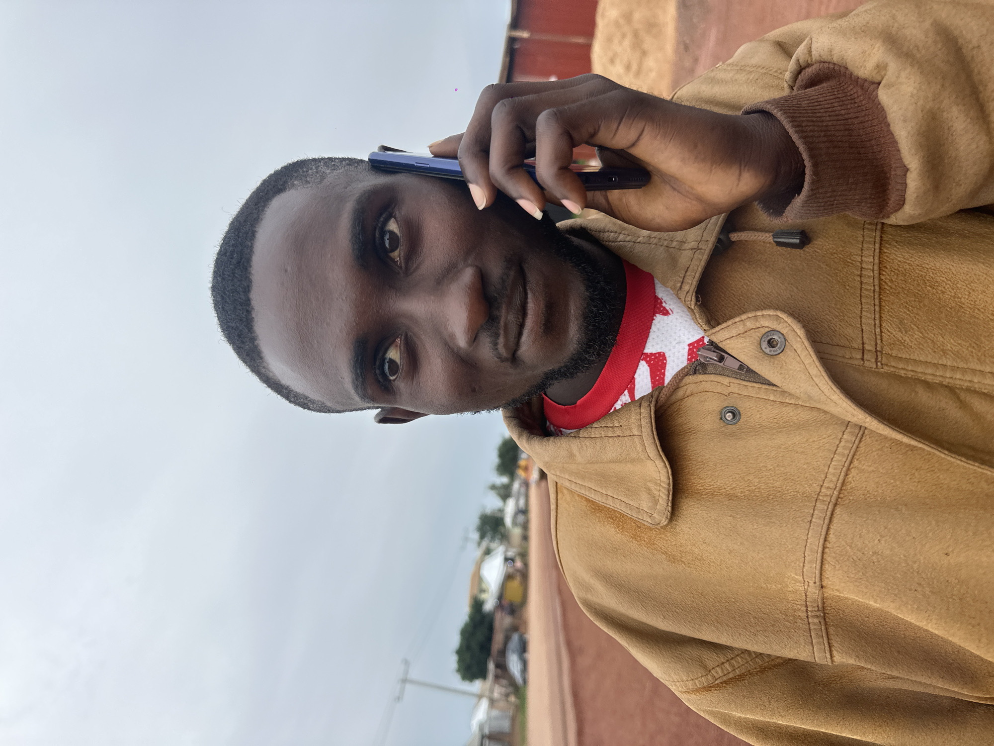A medium shot captures a Black man in his late 20s or early 30s holding a dark-colored smartphone to his right ear and speaking, with his gaze directed upwards and to his left. He is wearing a brown, collared jacket over a red and white patterned shirt. The jacket is zipped up, and a metal button is visible on the left lapel. The man has short, dark hair and a short beard.
The background suggests an outdoor, rural or semi-urban setting. A dirt road is prominent in the foreground and extends into the middle ground. To the left of the road, there are several buildings, some appearing to be small shops or homes with corrugated metal roofs, as well as a utility pole and some trees. A dark-colored car is parked on the side of the road. To the right of the man, there is a large, red structure, possibly a building or wall, and a pile of what appears to be hay or straw. The sky is overcast with light gray clouds. The overall impression is a scene in the city of Durumi, Nigeria, possibly on the outskirts or in a less developed area.