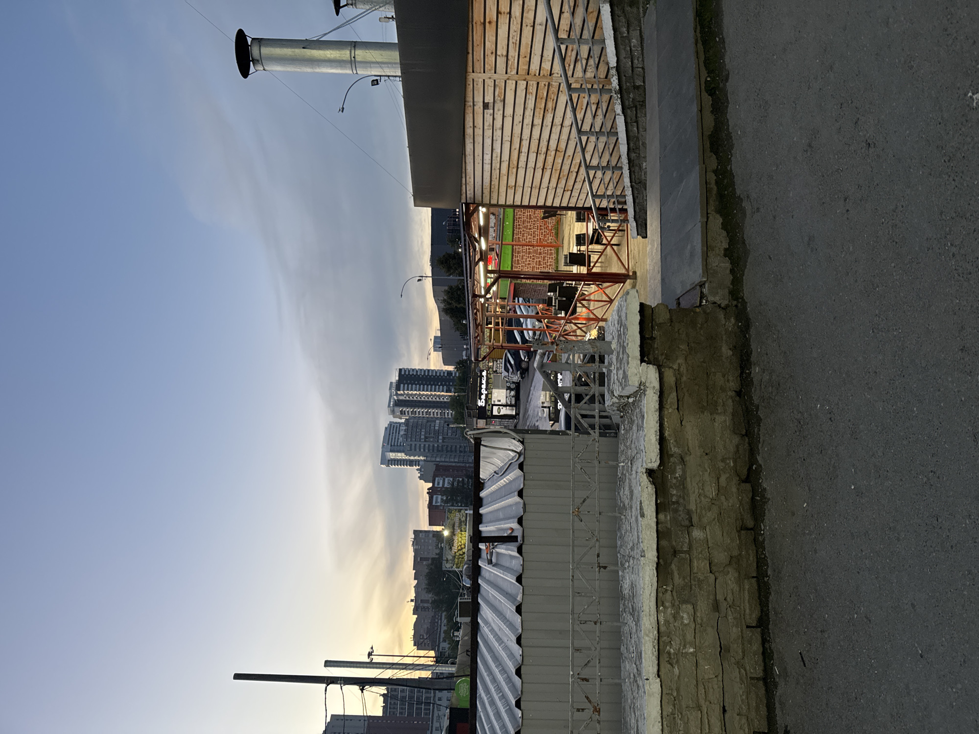 The image shows an exterior view of a restaurant with an outdoor seating area and a view of a city skyline at dusk. The restaurant features a wooden-clad building with a large metal chimney. The outdoor seating area is enclosed by a metal railing and has tables and chairs. In the background, there are several high-rise buildings, suggesting an urban setting. The sky is overcast with hints of sunset colors. The foreground shows a paved area with a low stone wall. The location is identified as Yekaterinburg, Russia. There are no people visible in the image.