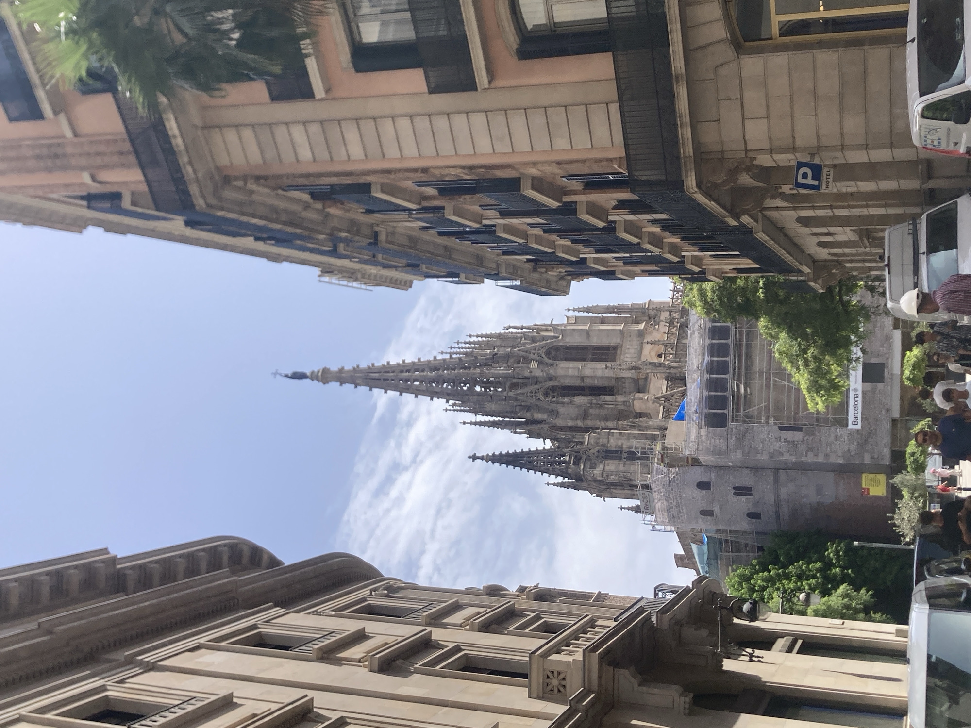 The image depicts a street view in Barcelona, Spain, with the Barcelona Cathedral's Gothic spires prominently featured in the background. On the left, a light-colored building with multiple windows and decorative elements lines the street. On the right, a salmon-colored building with dark balconies and windows stands tall. Scaffolding and construction materials are visible around a lower section of the cathedral, suggesting ongoing work. Several trees add greenery to the scene. In the foreground, people are walking on the sidewalk, and vehicles, including a white van and a car, are parked on the street. A blue parking sign with a "P" and "HOTEL" text is affixed to the salmon-colored building. The sky is clear with some light clouds.