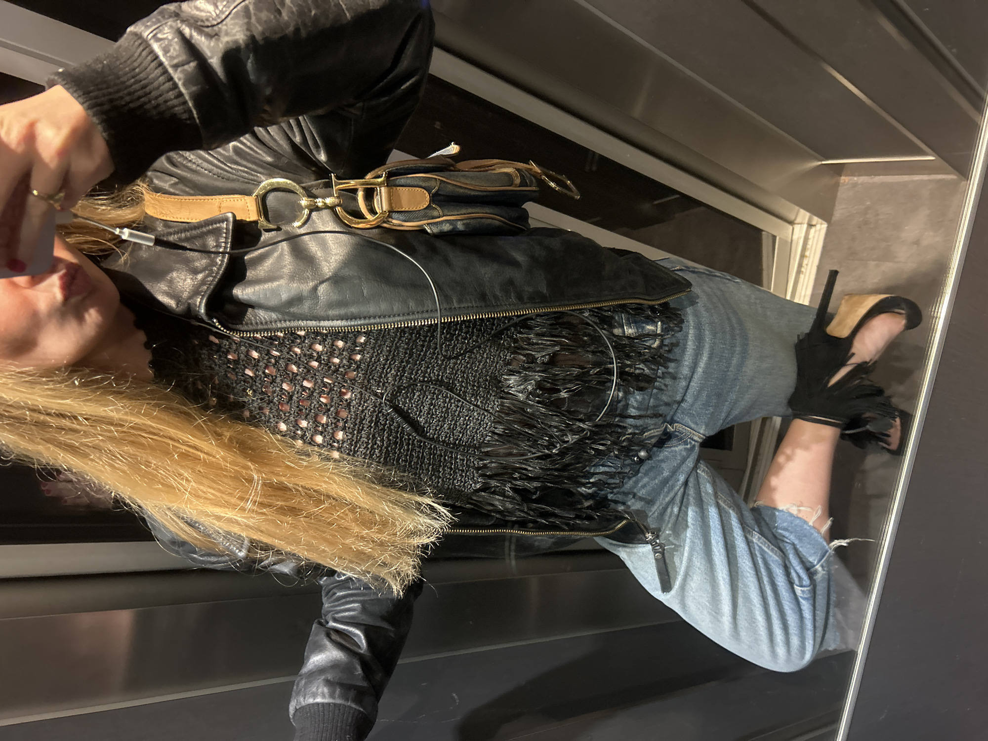 A person is pictured in a room that appears to be indoors, possibly a dressing room or a walk-in closet. They are wearing a black leather jacket over a black, fringed top, paired with light-wash jeans that have a ripped detail at the hem. A tan belt with a gold buckle is around the waist over the jacket, and a handbag is placed on top of the jacket near the belt. The person is also wearing black high-heeled sandals with fringe details. The background consists of what looks like stainless steel surfaces and a neutral wall, which contributes to an enclosed and perhaps modern setting. The image is taken from an unusual angle, suggesting a mirror reflection or the person is looking down at the camera. The location is in an unidentified city (Unknown, Unknown).