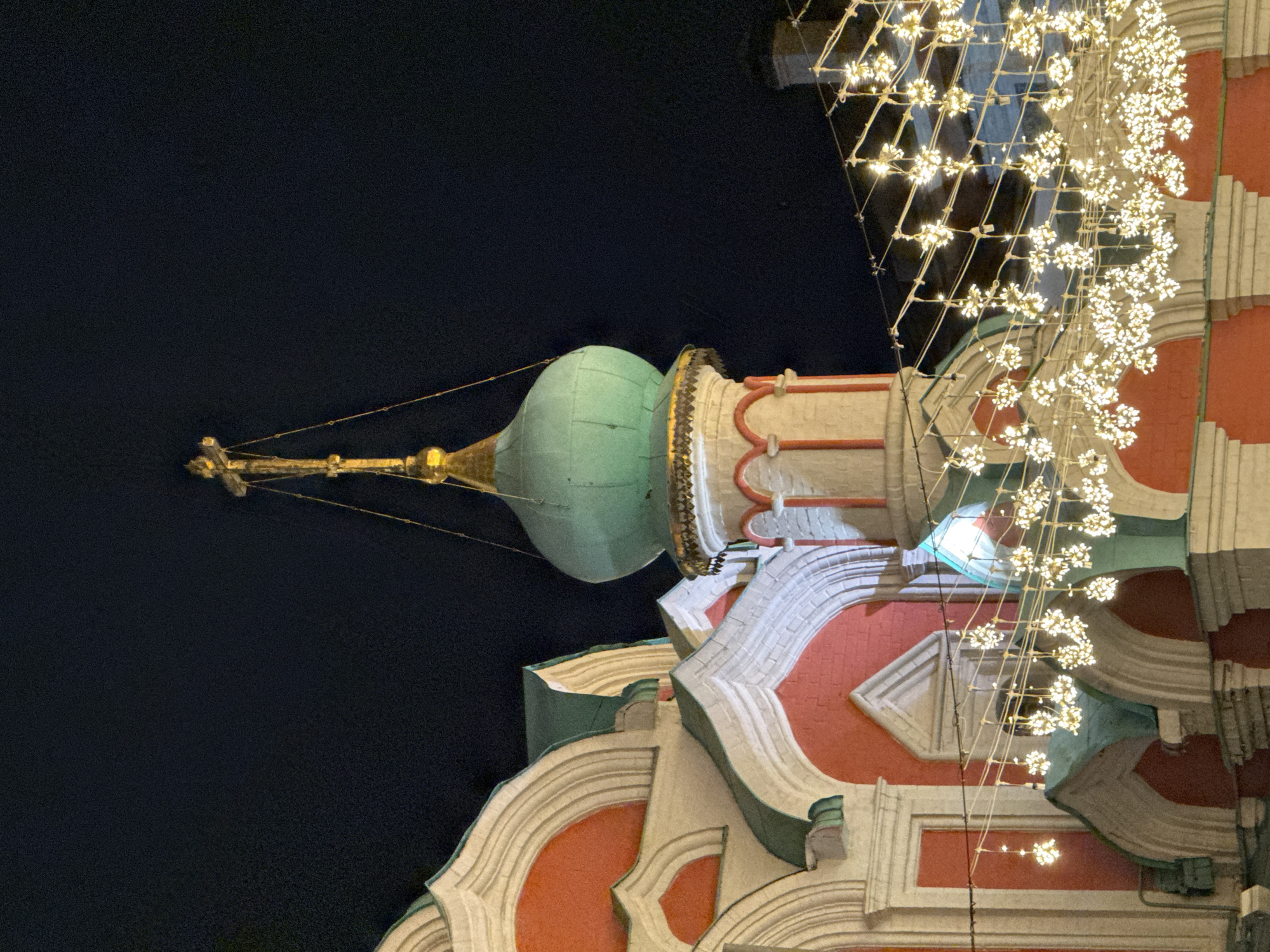The image shows a section of the St. Basil's Cathedral in Moscow, Russia at night. The angle is looking up at one of the building's onion-shaped domes and the decorated facade. String lights hang adjacent to the building. The background is a dark, featureless night sky.