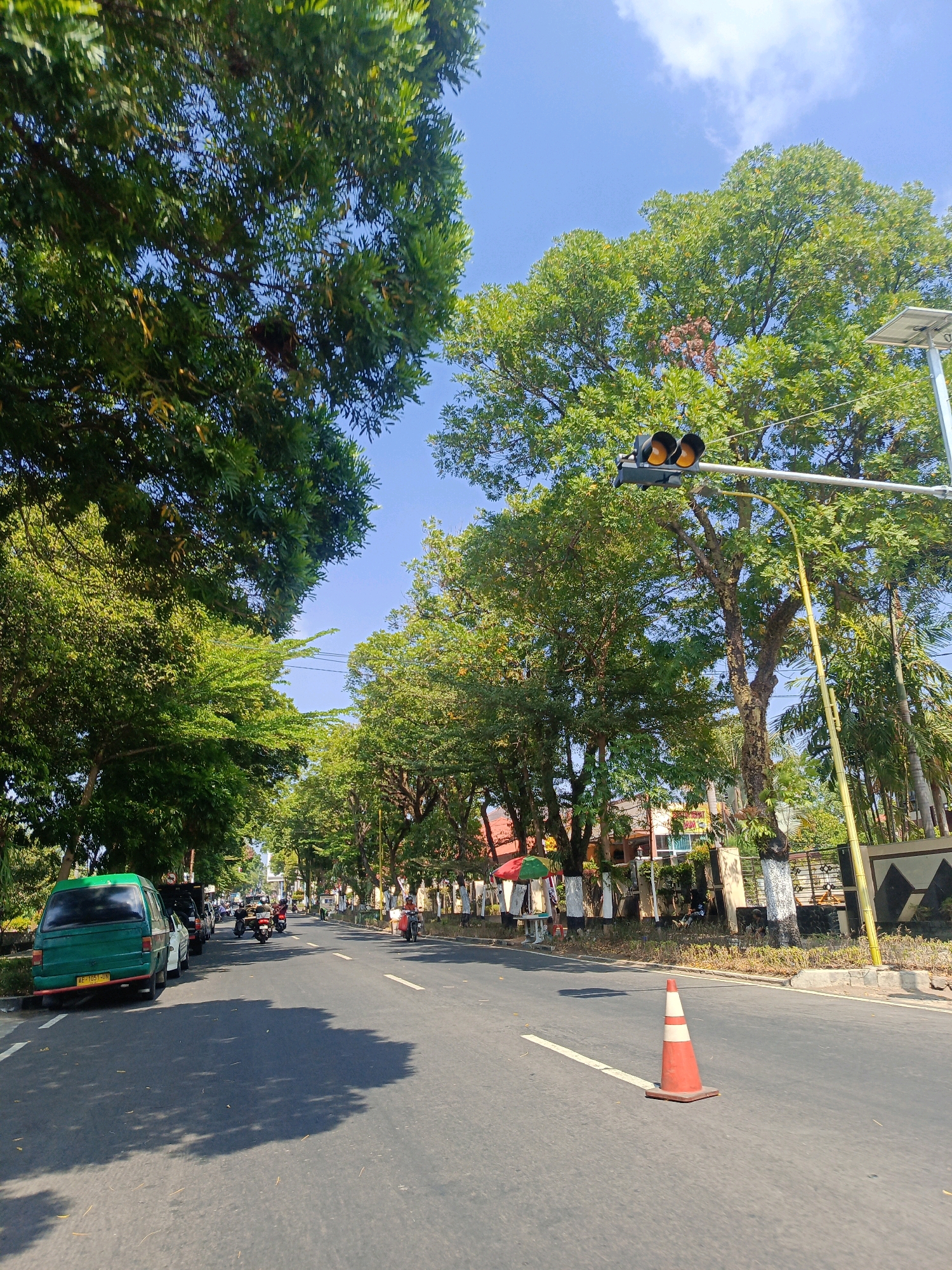 The image captures a street scene in Tambran, Indonesia, under a clear blue sky with scattered clouds. A road curves slightly to the right, bordered by lush green trees on both sides, creating a shaded canopy effect. In the foreground, a bright orange and white traffic cone is placed on the asphalt, near a white lane marker. A green van is parked on the left side of the road, followed by other vehicles and motorcycles. Further down the road, a traffic light with amber lights illuminated is visible, suspended over the intersection. Buildings and more trees line the right side of the street, with decorative elements on some tree trunks. The overall impression is of a well-maintained, tree-lined urban street on a sunny day.