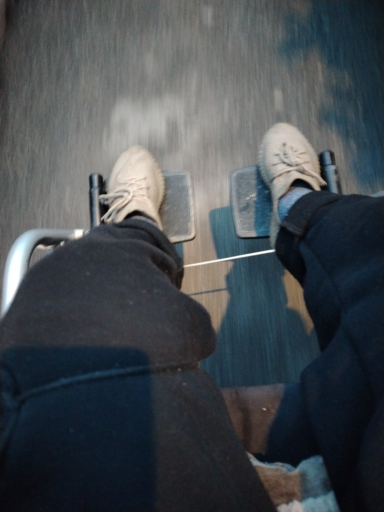 The image is an overhead, first-person perspective shot of someone sitting in a wheelchair. The subjects are the person's legs and feet, clad in light-colored sneakers and dark trousers. The wheelchair's footrests, which are textured and dark, are visible beneath the feet. The background is a blurry, streaky surface, suggesting movement, likely pavement or a similar outdoor ground. The lighting is dim, casting shadows and highlighting the texture of the ground and the person's clothing. The overall impression is one of motion, possibly a person being pushed or self-propelling in the wheelchair. There is no visible text. The scene appears to be set outdoors during daylight, though the dim lighting might suggest dusk or an overcast day.
