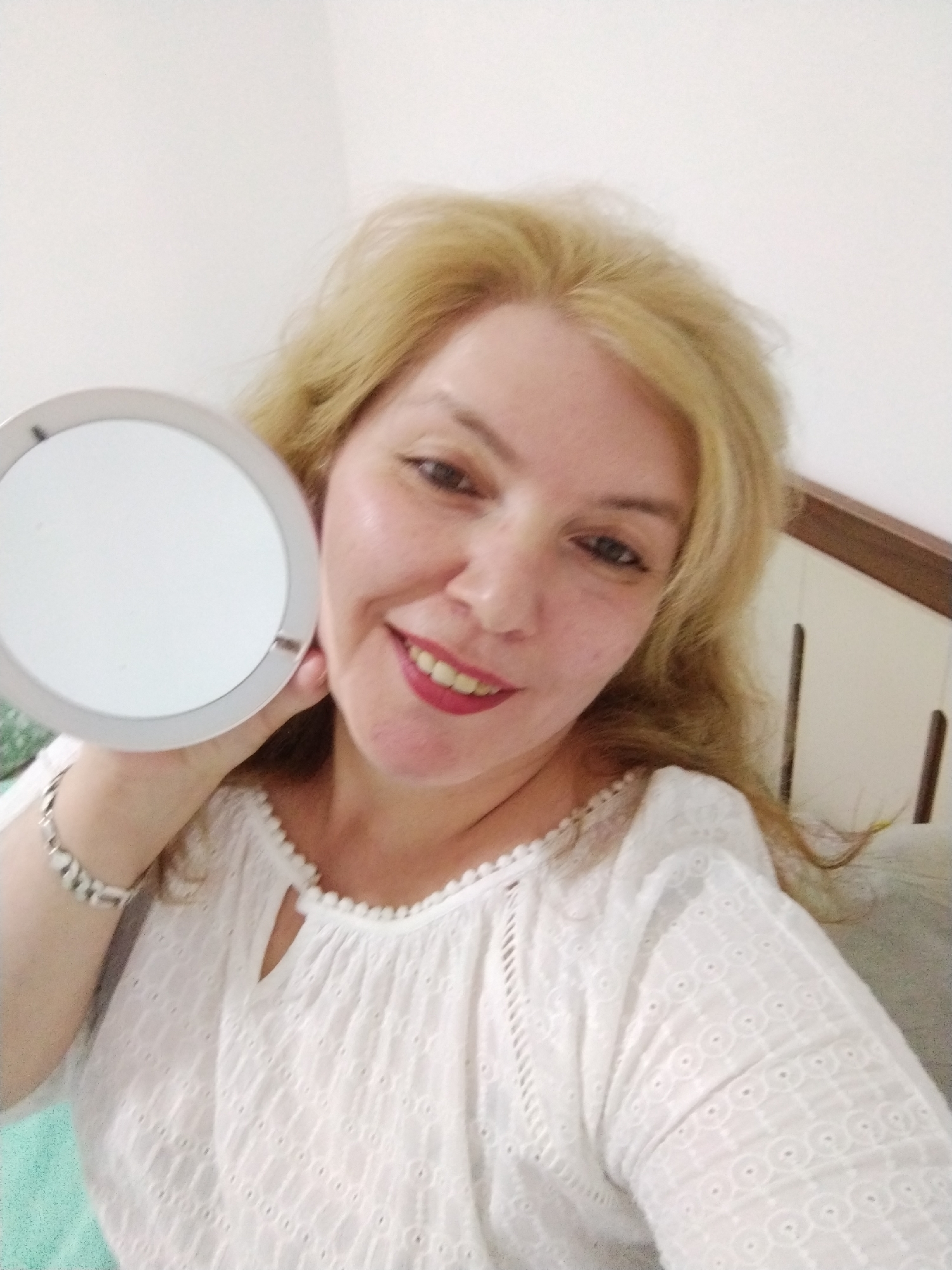 A woman with blonde hair and red lipstick smiles at the camera while holding a round mirror in her left hand. She is wearing a white, embroidered top with a keyhole neckline. A silver bracelet is visible on her left wrist. The background shows a white wall and a portion of a wooden headboard. The image appears to be taken indoors, likely in a residential setting. There is no indication that the location is Al Mas'oudi, United Arab Emirates.