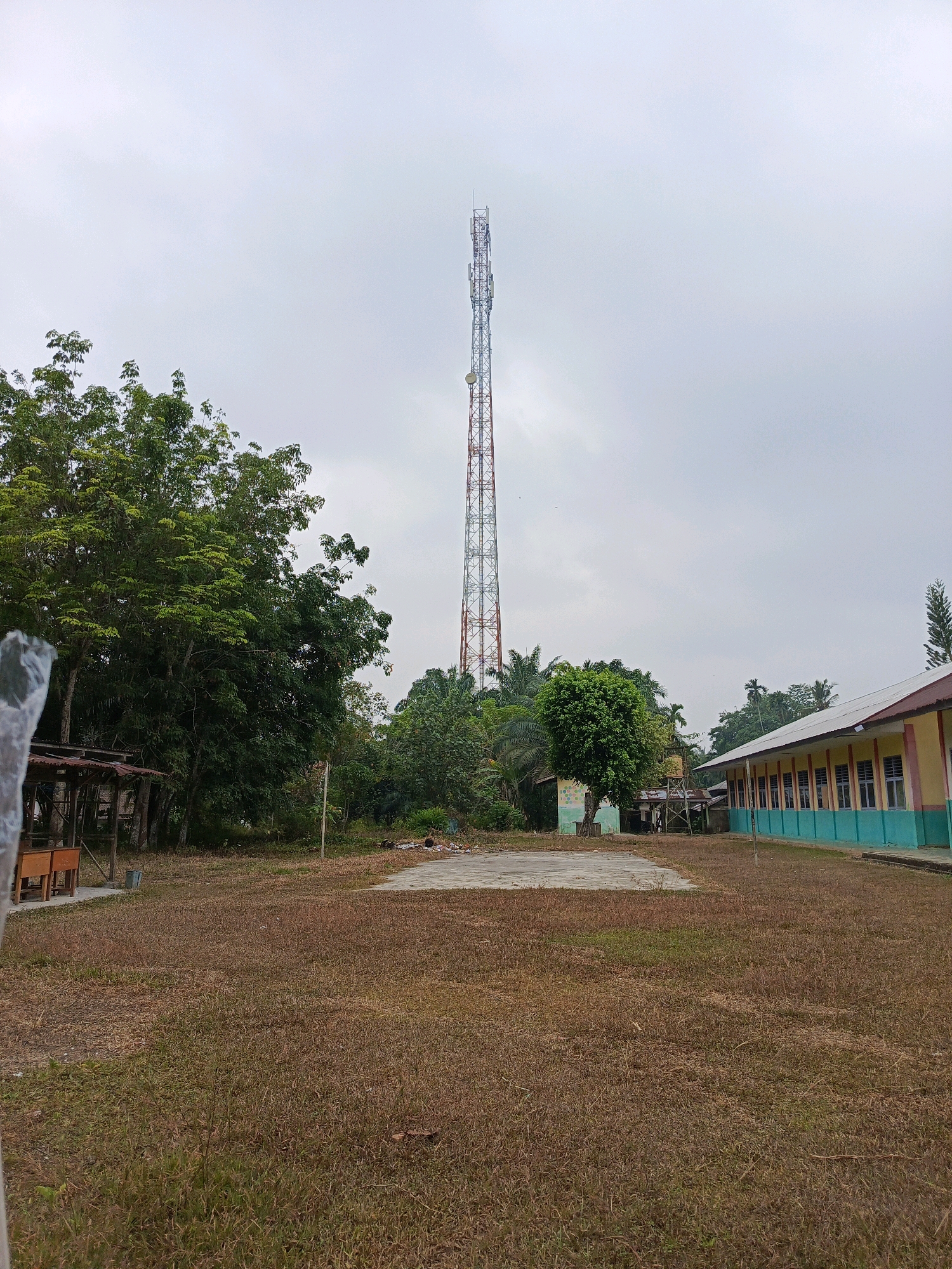 This image depicts an outdoor scene in what appears to be a rural or semi-rural area, possibly near a school in Wonosari, Indonesia. In the background, a tall telecommunications tower with red and white lattice structure dominates the sky. To the right of the tower is a low-lying building with a yellow facade, turquoise trim, and a white roof. The building has a row of windows with visible panes and frames. In the mid-ground, there is dense green foliage from trees and palm trees. In the foreground, a dry, brown grassy field stretches out, with a concrete rectangular area in the center, possibly a court or foundation. To the left of the concrete area, a simple wooden shelter with a corrugated roof is visible, along with some tables and chairs. The overall environment suggests a somewhat overcast day. There are no people or animals visible in the frame.