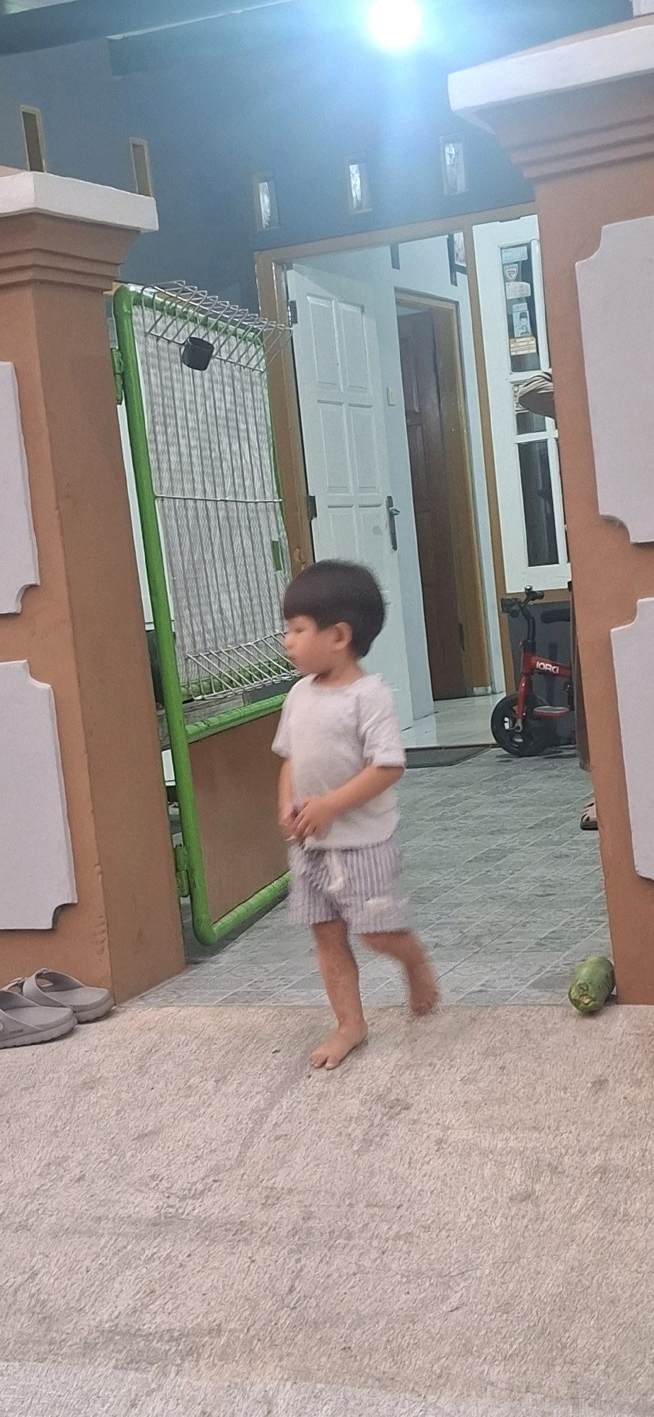 A young boy with dark hair, wearing a light-colored t-shirt and striped shorts, walks barefoot across a textured light-colored floor. He is in motion, with his right leg extended and his left leg bent. To his left, a pair of gray sandals are visible on the same floor surface. In the background, a green metal rack with a wire grid stands vertically. Beyond the rack, a doorway leads into a building with a white door. Further in the background, a red child's bicycle with the word "IORGI" on it is propped up. To the right of the boy, a green, oblong object rests on the floor. The scene appears to be outdoors, possibly a porch or pathway, with a wall to the immediate left and right of the frame, painted in earthy tones. The lighting suggests it is evening or dusk, with a bright light source visible at the top of the frame. The general environment is consistent with a residential area in Desa Gagaksipat, Indonesia.