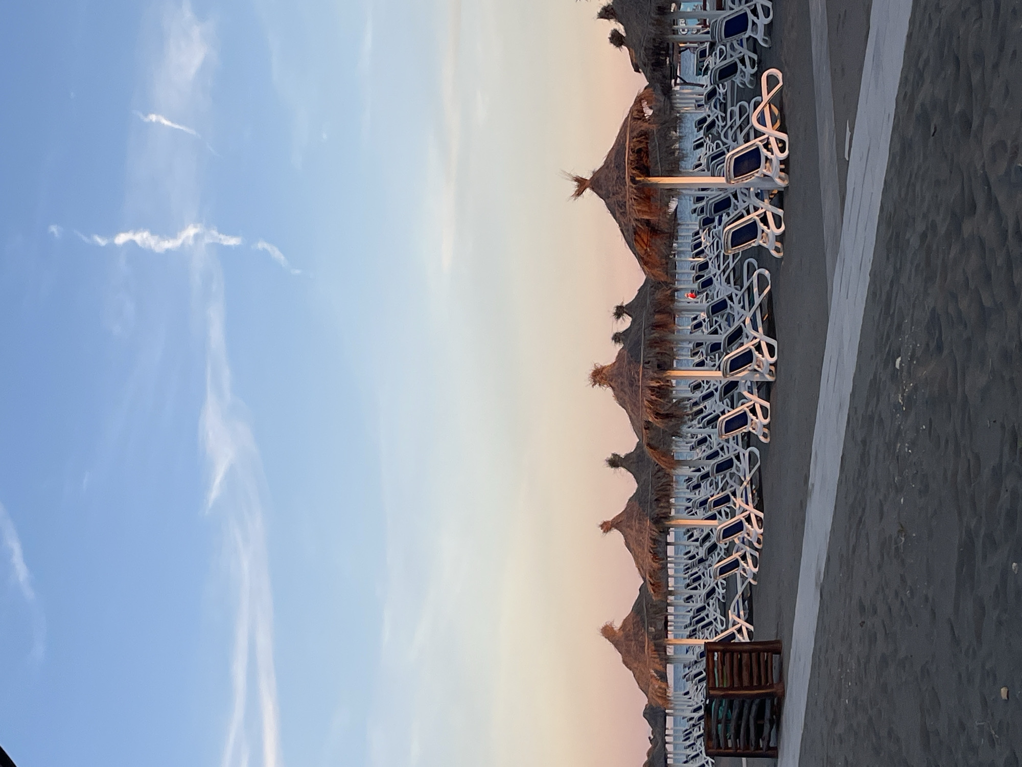 A wide shot captures a tranquil beach scene at dusk in Pistula, Montenegro. Rows of white lounge chairs with blue cushions are neatly arranged under thatched-roof umbrellas on the dark sand. The sky transitions from a pale blue at the top to a soft pink and orange near the horizon, indicating the setting sun. A few wispy clouds streak across the sky. In the foreground, the dark sand stretches towards the viewer, with a white path running horizontally across the scene. A small wooden structure, possibly a bin or signpost, is visible on the left side of the path. The ocean is a calm expanse of blue behind the rows of chairs and umbrellas. No people are actively engaged in any activity, and the overall impression is one of quiet repose.