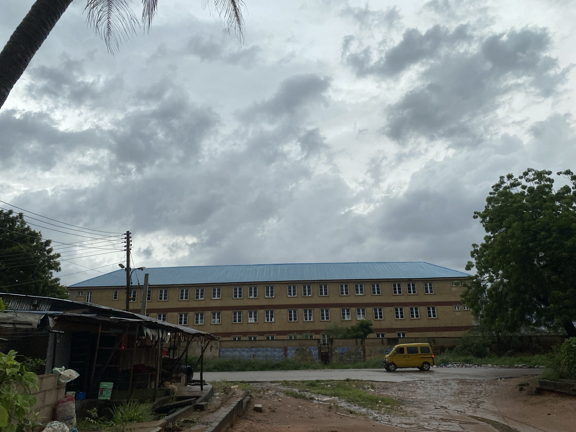 The image shows a street scene in Lagos, Nigeria under a cloudy sky. A three-story building with a blue roof and numerous windows dominates the background. A small, yellow vehicle is on the road in front of the building. In the foreground, there's a makeshift structure with a corrugated iron roof. Vegetation is present throughout, including a large tree on the right side of the frame and palm trees at the top. Utility poles and wires are visible. The road appears damp or wet. The location seems to be a less developed area, with some litter visible.