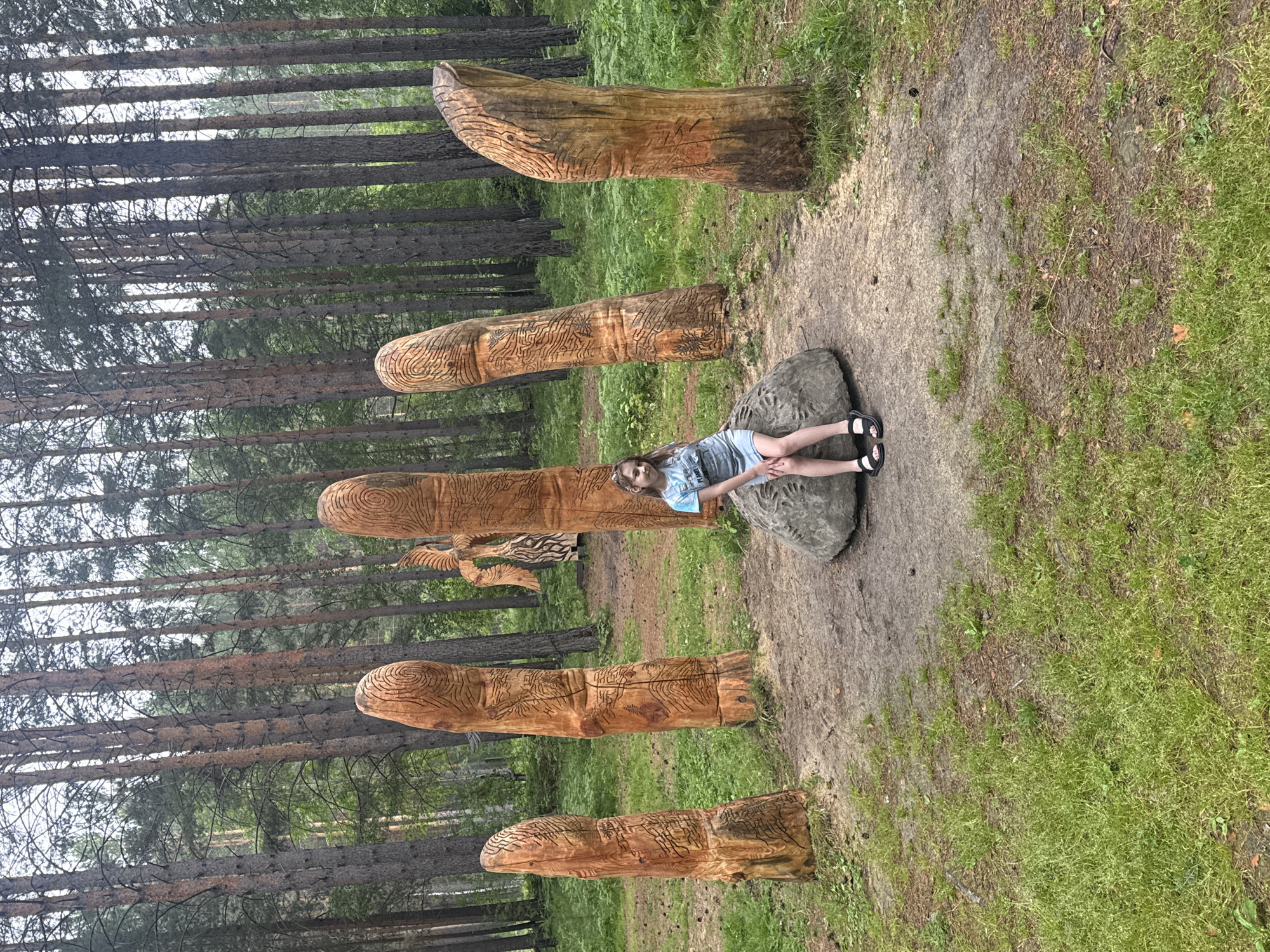 A young girl with blonde hair, wearing a light blue and white tie-dye t-shirt and a denim skirt, sits on a large gray rock in a forest clearing. She is smiling and looking at the camera. Around her are five large, intricately carved wooden sculptures that resemble fingers, with the patterns of fingerprints etched into the tops. Behind the girl and to the right, another wooden sculpture appears to be a stylized bird or winged figure. The clearing is surrounded by a dense forest of tall pine trees. The ground is covered in a mixture of dirt, pine needles, and green grass with some patches of ferns. The overall setting is a natural, outdoor art installation within a forest. This scene is located in Savvateevka, Russia.