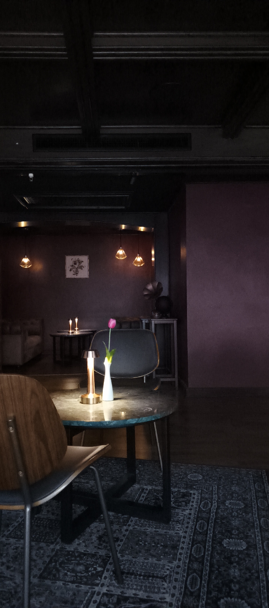 The image captures the interior of a dimly lit, stylish bar, lounge, or cafe, creating an intimate and cozy atmosphere. The scene is devoid of people.
In the immediate foreground, a round table with a dark, possibly mosaic or stone surface is central. On this table, a modern, cylindrical table lamp with a metallic copper or bronze finish casts a warm glow, illuminating a slender white vase holding a single pink tulip. Partially visible to the left is the back of a wooden chair with a dark seat.
The midground reveals more of the lounge area. Another table with two small, candle-like lights is situated further back. A dark, upholstered chair with a curved back is also visible, along with hints of a sofa or lounge seating on the left.
The background features a wall painted in a deep, muted purple or maroon hue. A framed picture with a floral design hangs on this wall. Above it, several pendant lights with geometric glass shades emit warm light. To the right, a vintage-style phonograph or gramophone with a large horn rests on a dark side table. The ceiling is dark, possibly black, with visible beams and air vents. A small security camera can be discerned in the upper left area of the ceiling.
The floor is covered by a dark, intricately patterned rug, with darker wooden flooring visible in some areas. The overall impression is one of a relaxed, evening setting, designed for quiet socializing or unwinding. The time of day is clearly night or evening, given the reliance on artificial light sources. No text is visible in the image. The mood evoked is calm, sophisticated, and inviting.